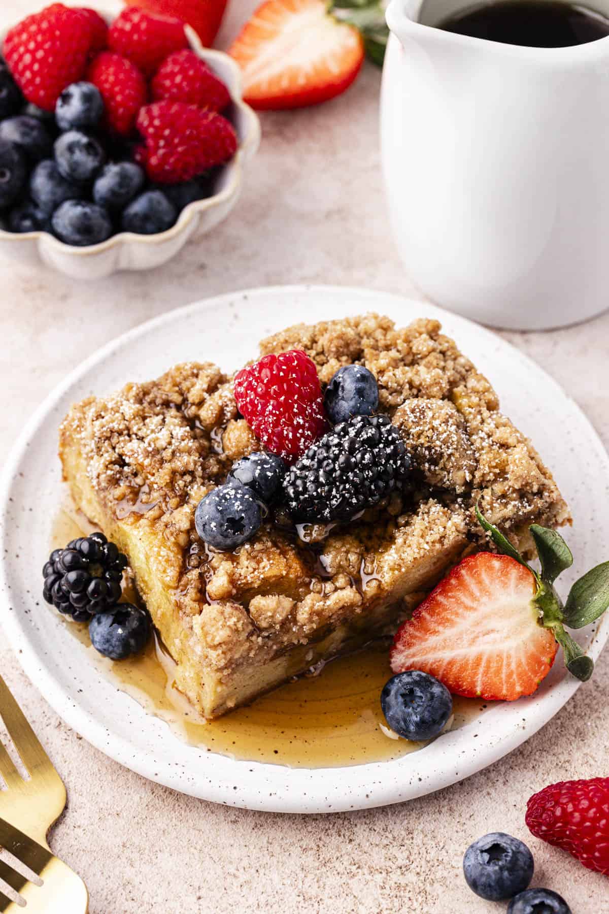 a small plate with a slice of french toast casserole on it topped with maple syrup, fresh berries and a light dusting of powered sugar beside a small bowl of fresh berries, a white dish of syrup, two forks and more berries scattered around