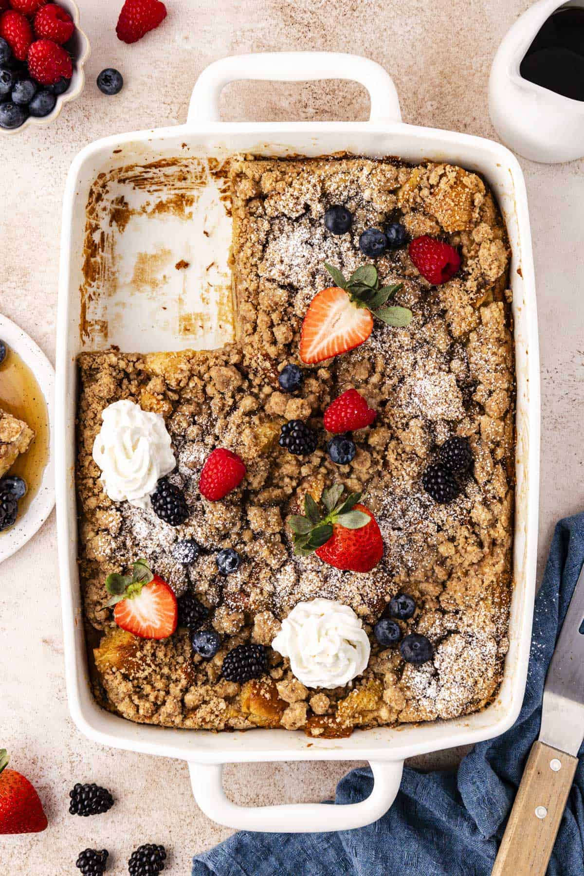 over head view of a white baking dish full of french toast casserole with one slice missing, topped with fresh berries, dollops of whipped cream and a light dusting of powdered sugar, surrounded by a bowl of berries, a dish of syrup, a plate with a slice and syrup on it, a blue kitchen towel and a spatula