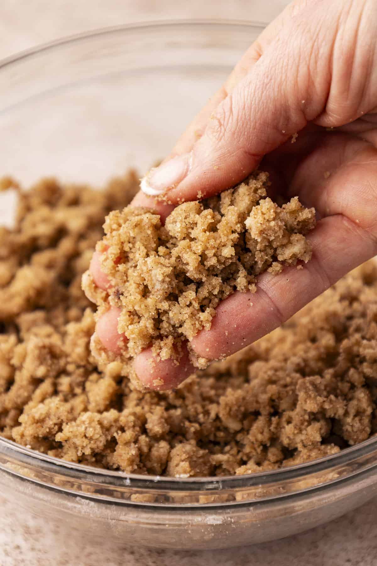 a hand holding crumb topping over top of a clear glass bowl full of more crumb topping