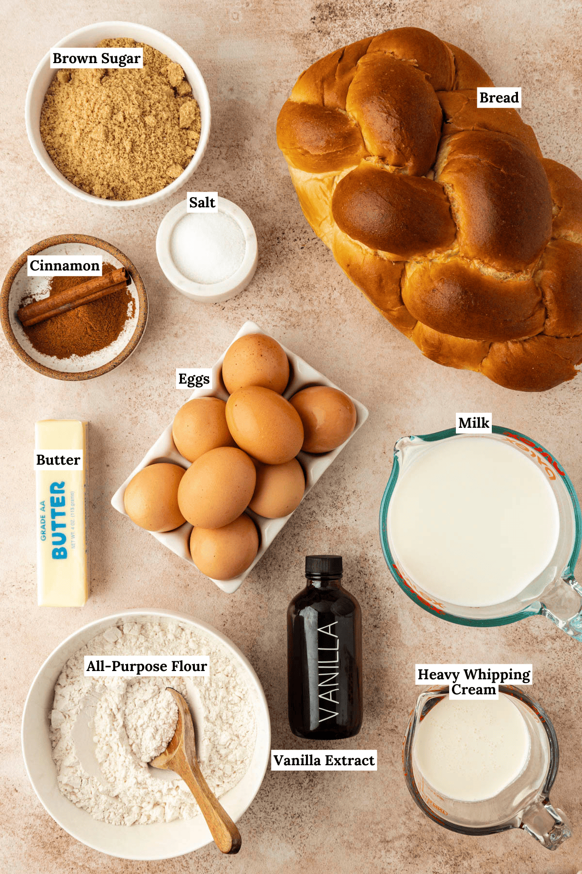 over head view of the ingredients for french toast casserole including a loaf of bread, a bowl of brown sugar, a small bowl of salt, a bowl of cinnamon, eight brown eggs, a stick of butter, a measuring cup of milk, a measuring cup of heavy whipping cream, a bottle of vanilla extract and a bowl of all-purpose flour