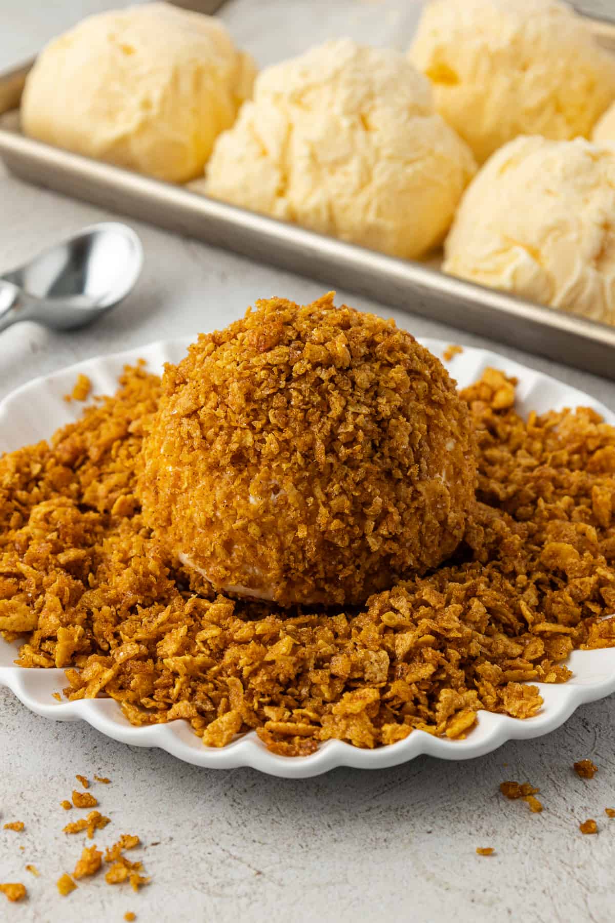 a ball of ice cream coated in a corn flake cereal mixture on top of more crushed corn flake mixture on a white plate, beside a sheet pan with rows of vanilla ice cream balls on it