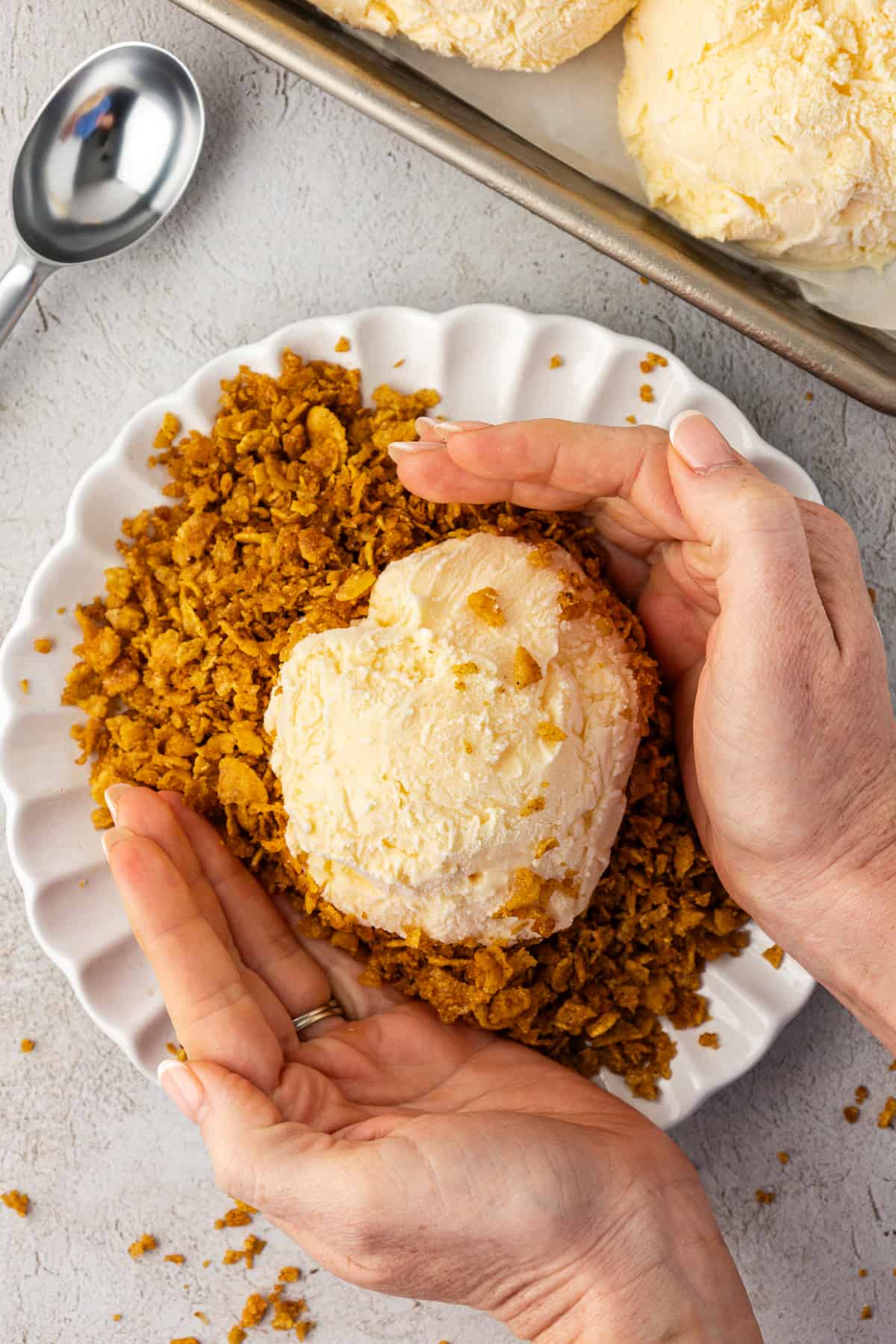 two hands pressing a corn flake cereal mixture over a ball of vanilla ice cream on a white plate beside a sheet pan of vanilla ice cream balls