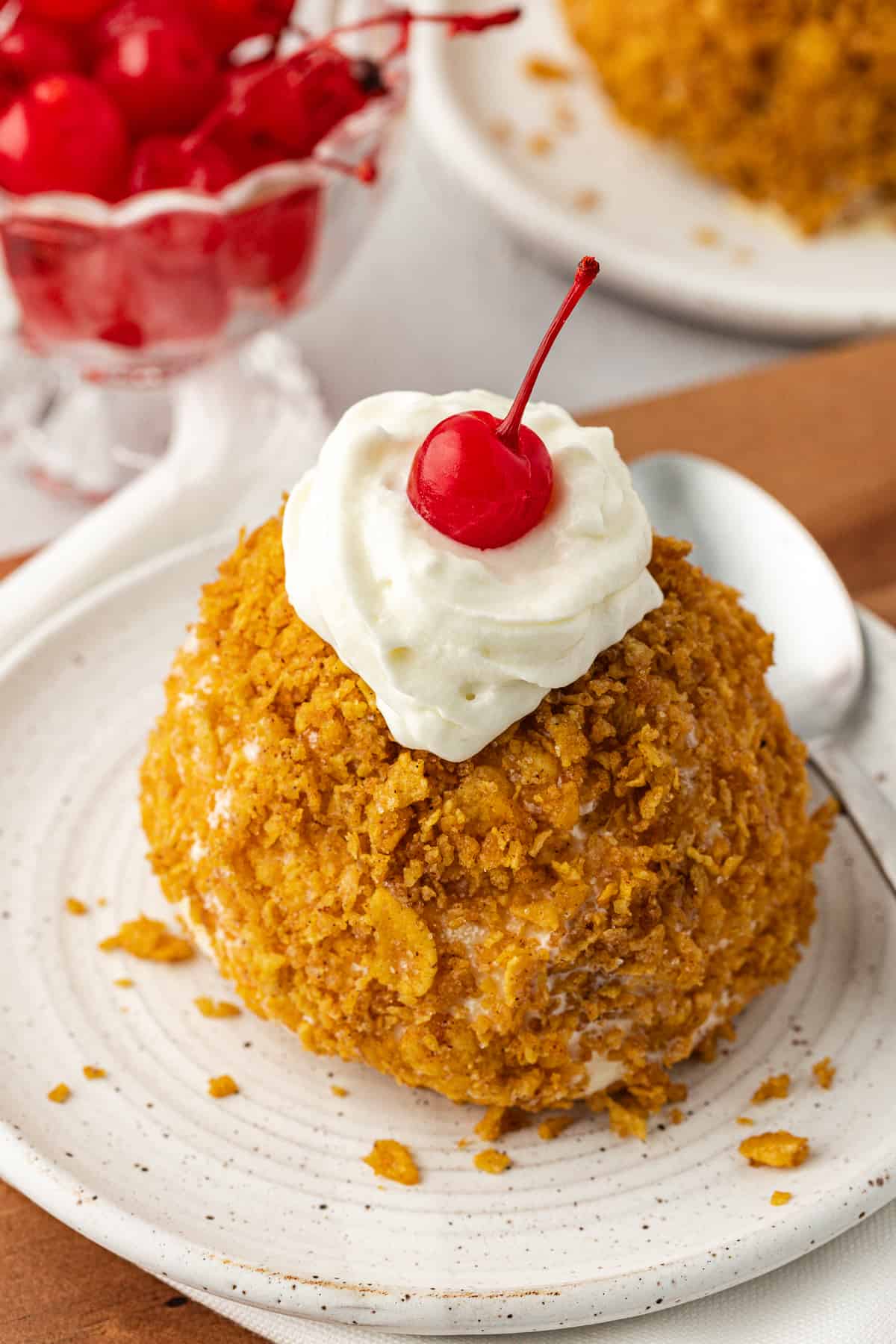 a ball of fried ice cream on a small plate with a spoon, topped with whipped cream and a cherry with a bowl of cherries and more fried ice cream in the background