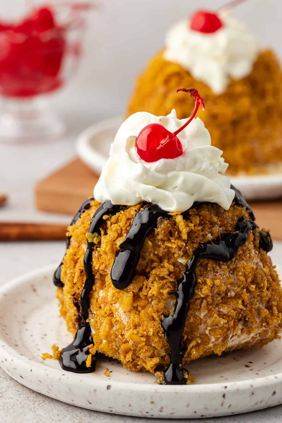 fried ice cream topping with chocolate syrup, whipped cream and a cherry on a small plate with another plate of fried ice cream and a bowl of cherries in the background