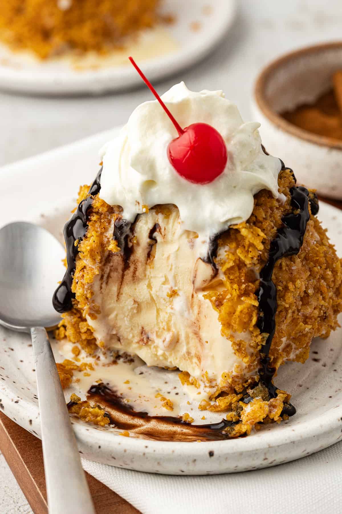 a ball of fried ice cream topping with chocolate syrup, whipped cream and a cherry on a small plate with a spoon and a bite missing from the ice cream