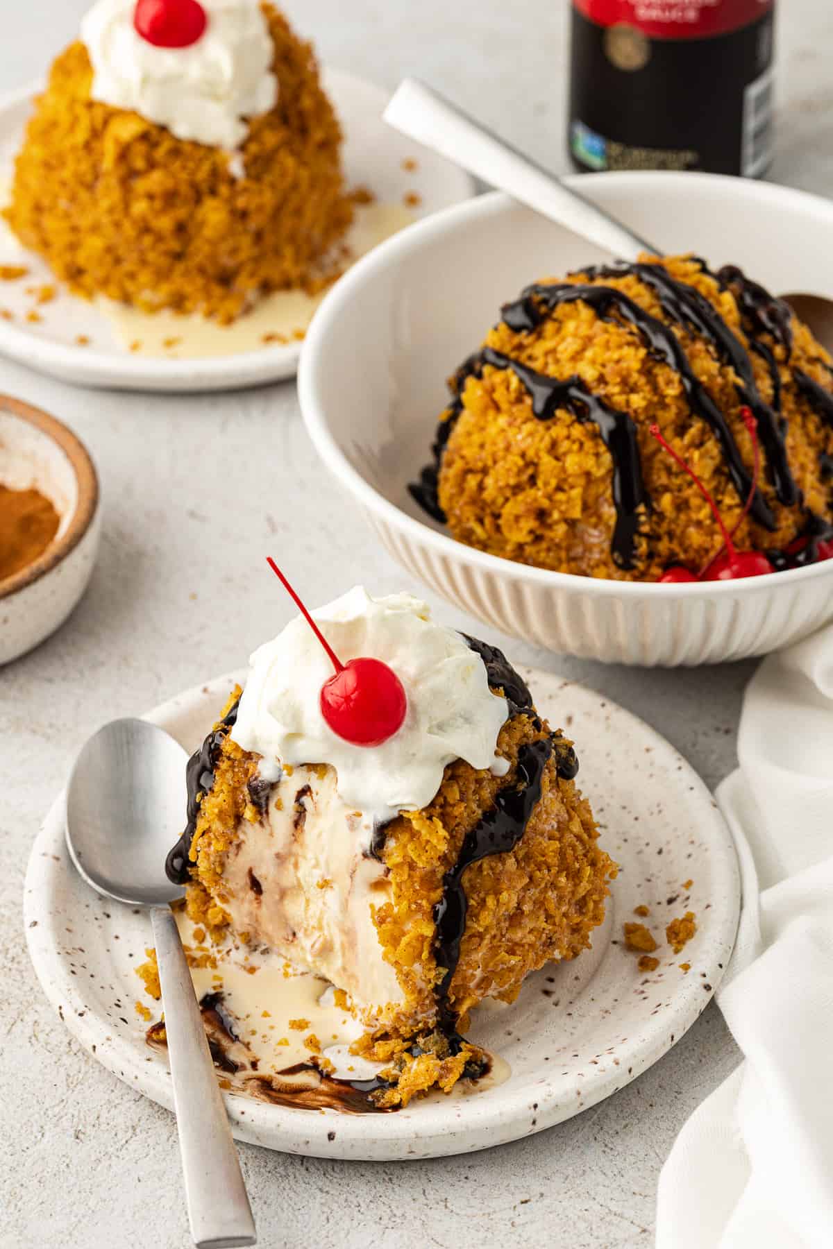 a small plate with fried ice cream partially eaten on it with a spoon, beside a bowl with another ball of fried ice cream, a white towel, and a plate with another ice cream ball in the background