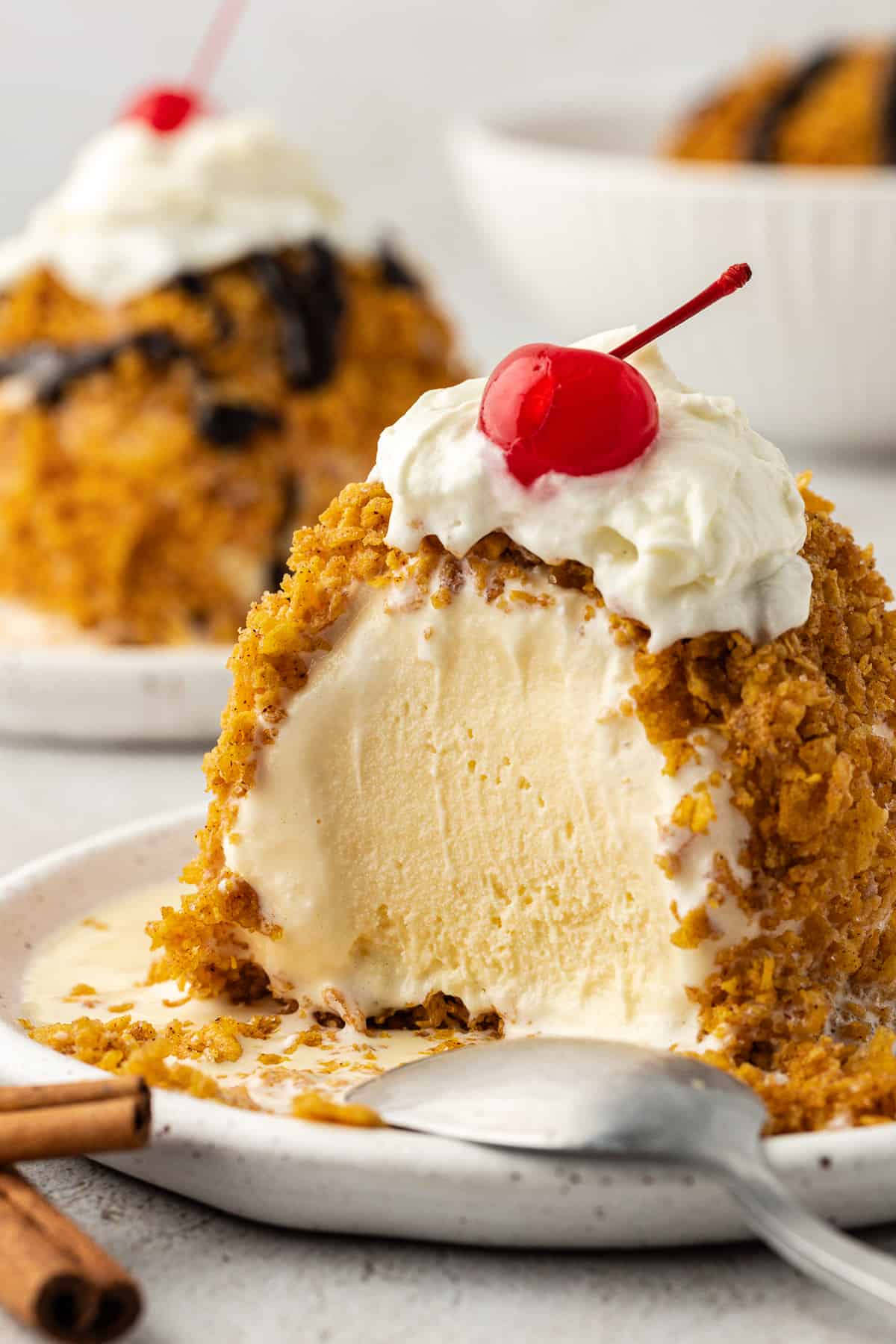 fried ice cream on a plate with a spoon leaning on the side of it with a bite missing, whipped cream and a cherry on top and another plate and a bowl of ice cream in the background