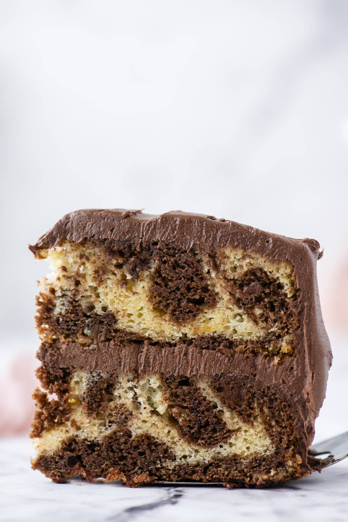 side view of a slice of marble cake on a spatula showing the marble effect on the inside of the cake and the layer of chocolate frosting in between the two layers of cake