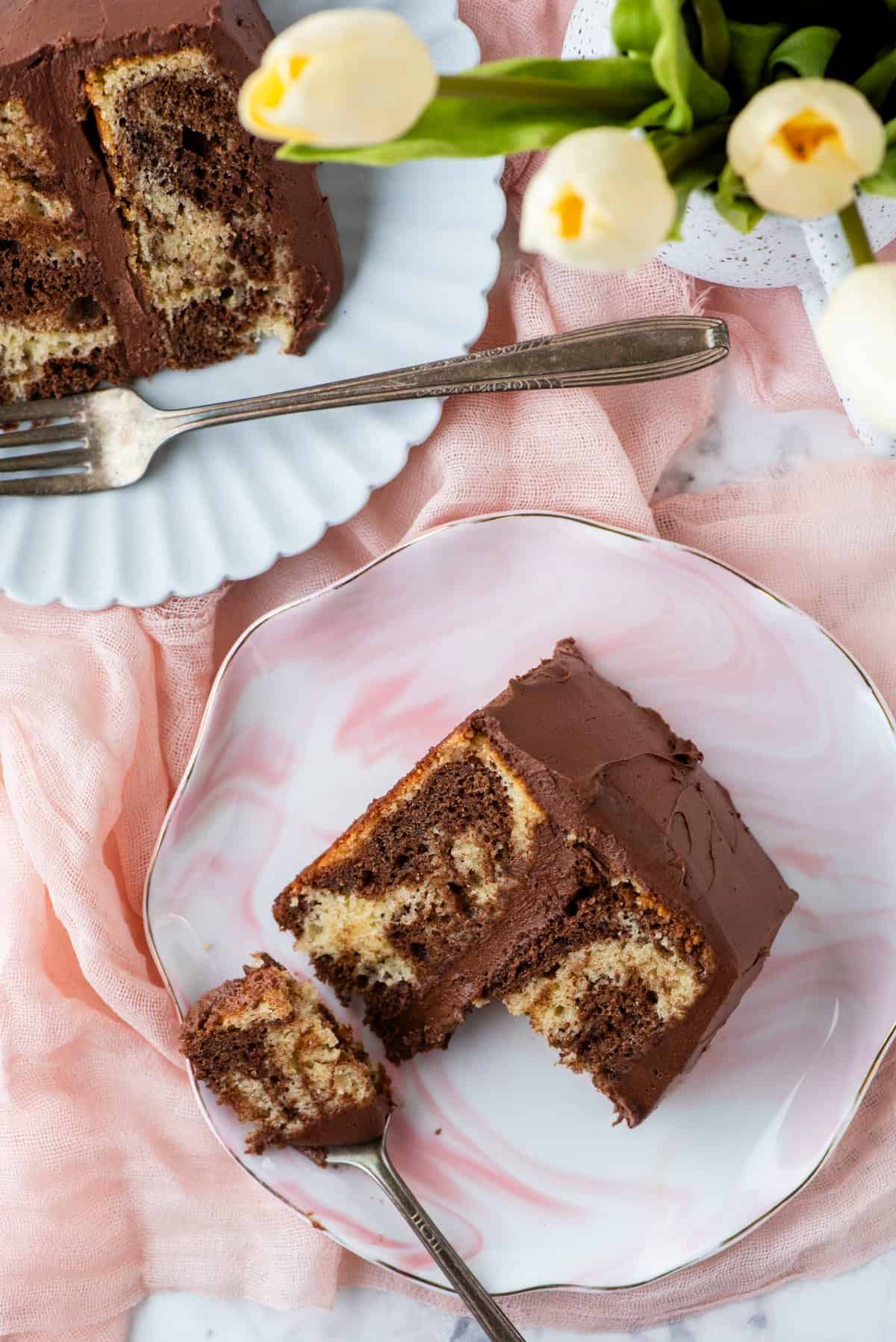 overhead view of two slices of marble cake on two small plates with forks on top of a light pink cloth with white tulips beside them