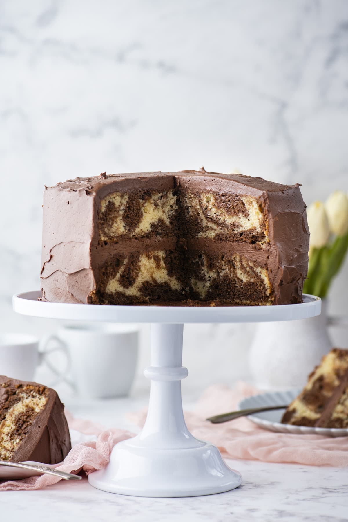 a two layer swirl cake with chocolate frosting on a white cake stand with two slices missing that are sitting on small plates with forks on a light pink cloth near the cake stand