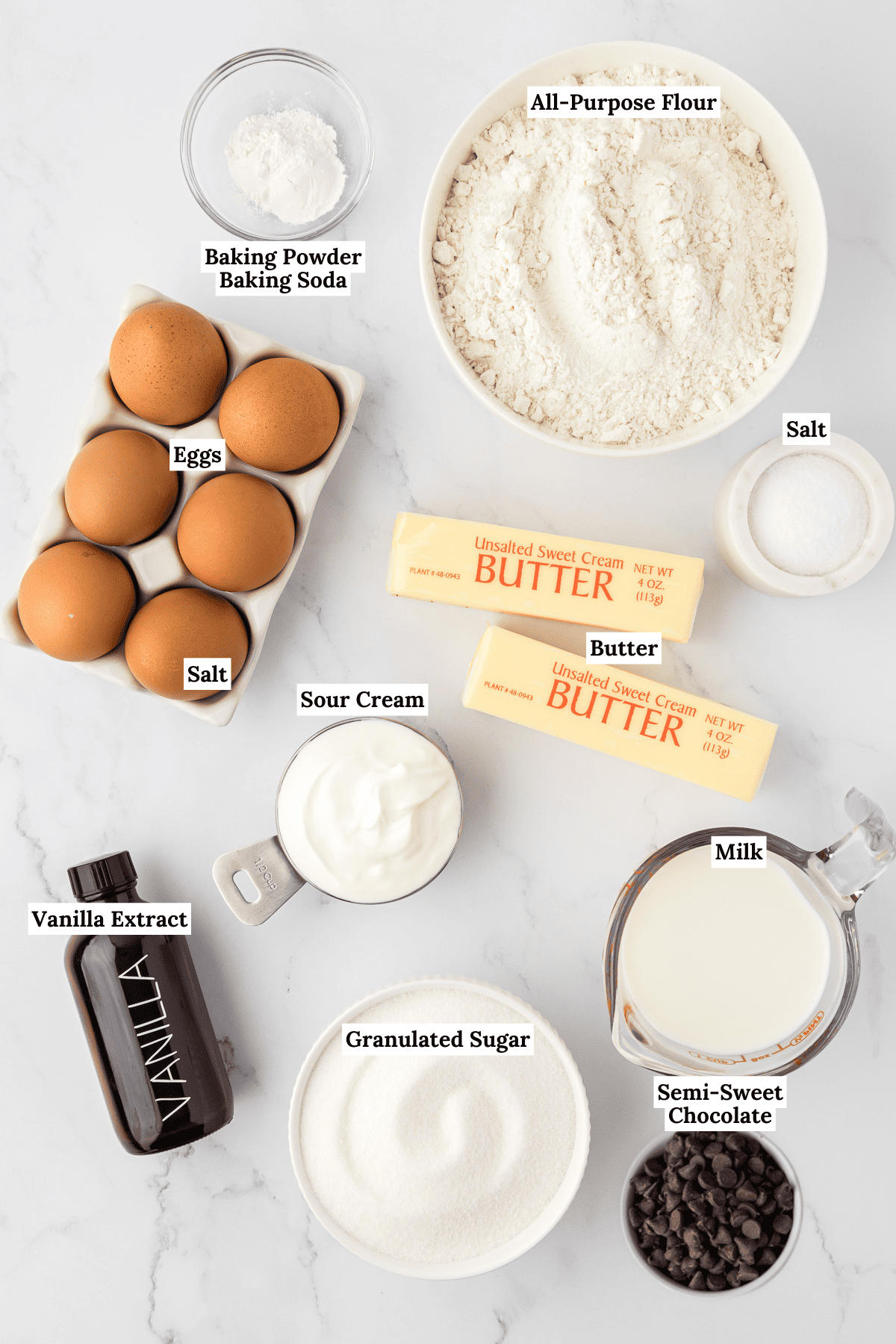 overhead view of the ingredients for marble cake including baking powder, baking soda, all-purpose flour, salt, two sticks of butter, 6 eggs, sour cream, milk, granulated sugar, vanilla extract and semi-sweet chocolate chips