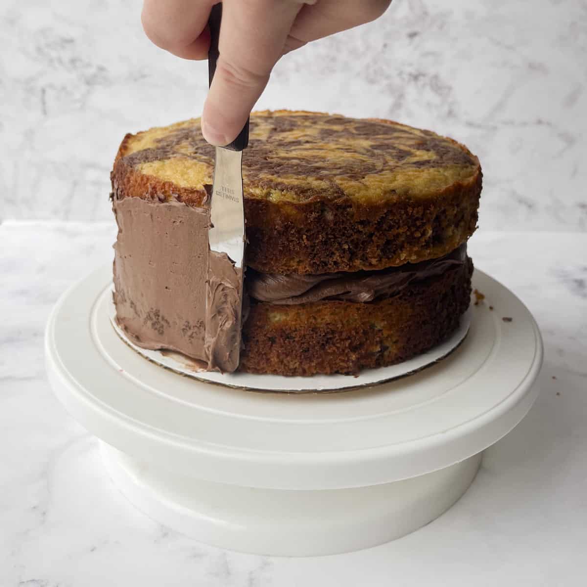 two layers of cake with chocolate frosting in between, sitting on a cardboard round on top of a white cake stand, being frosted on the side with chocolate frosting