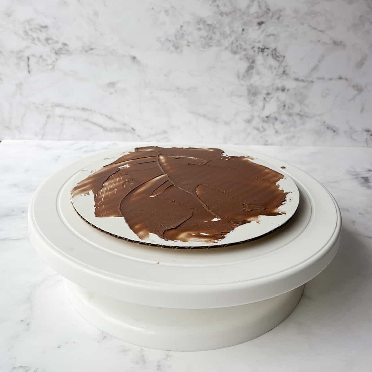 a white cake stand with a white cardboard round on it with a layer on chocolate frosting smeared on it