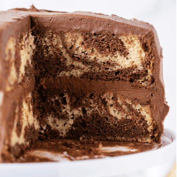 marble cake on a white cake stand with a large slice missing out of it, exposing the inside with the marble effect and two layers of cake with chocolate frosting in between