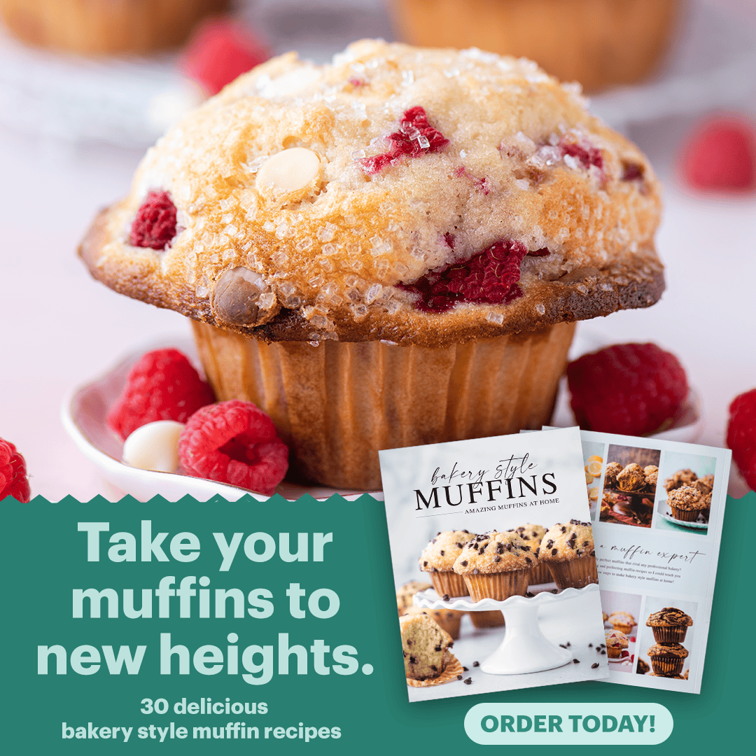 A close-up of a golden muffin with white chocolate chips and raspberries, surrounded by fresh raspberries. In the corner, two recipe booklets are displayed with the text: Take your muffins to new heights. Order today!.