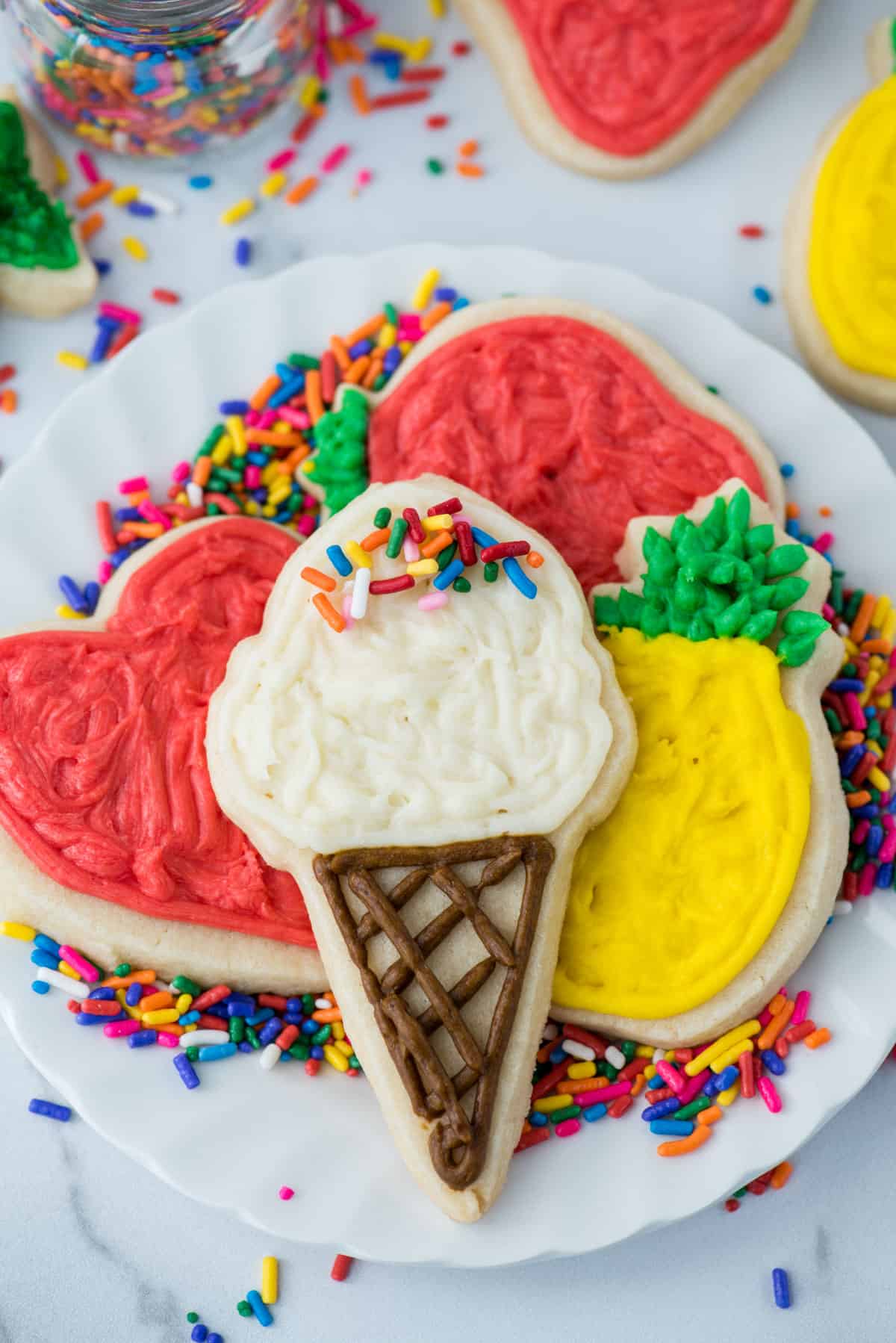 a white plate stacked with sugar cookies in different shapes with frosting decorations including an ice cream cone, a pineapple, and two hearts with piles of rainbow sprinkles underneath them and more sprinkles and cookies scattered around