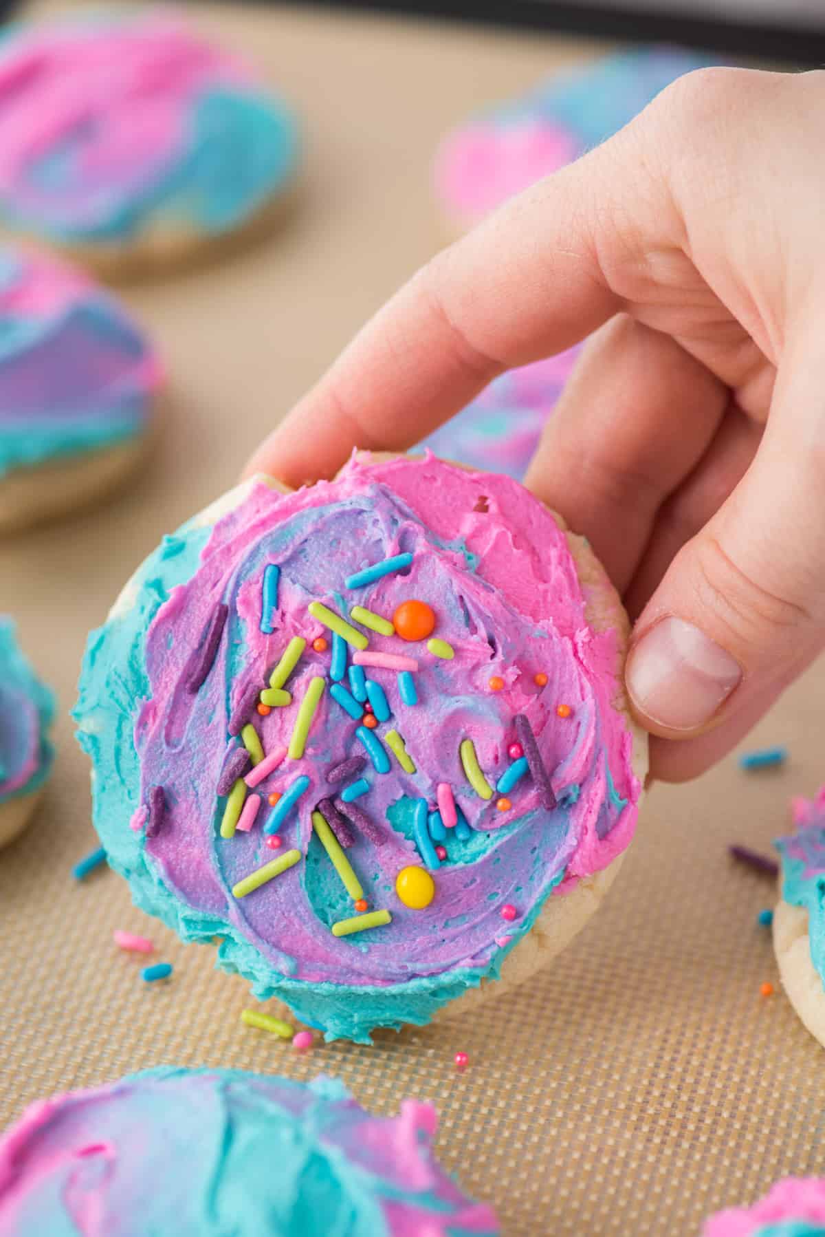 a sugar cookie decorated with a pink, purple and blue frosting and sprinkles being held up off a silicone mat with more cookies around