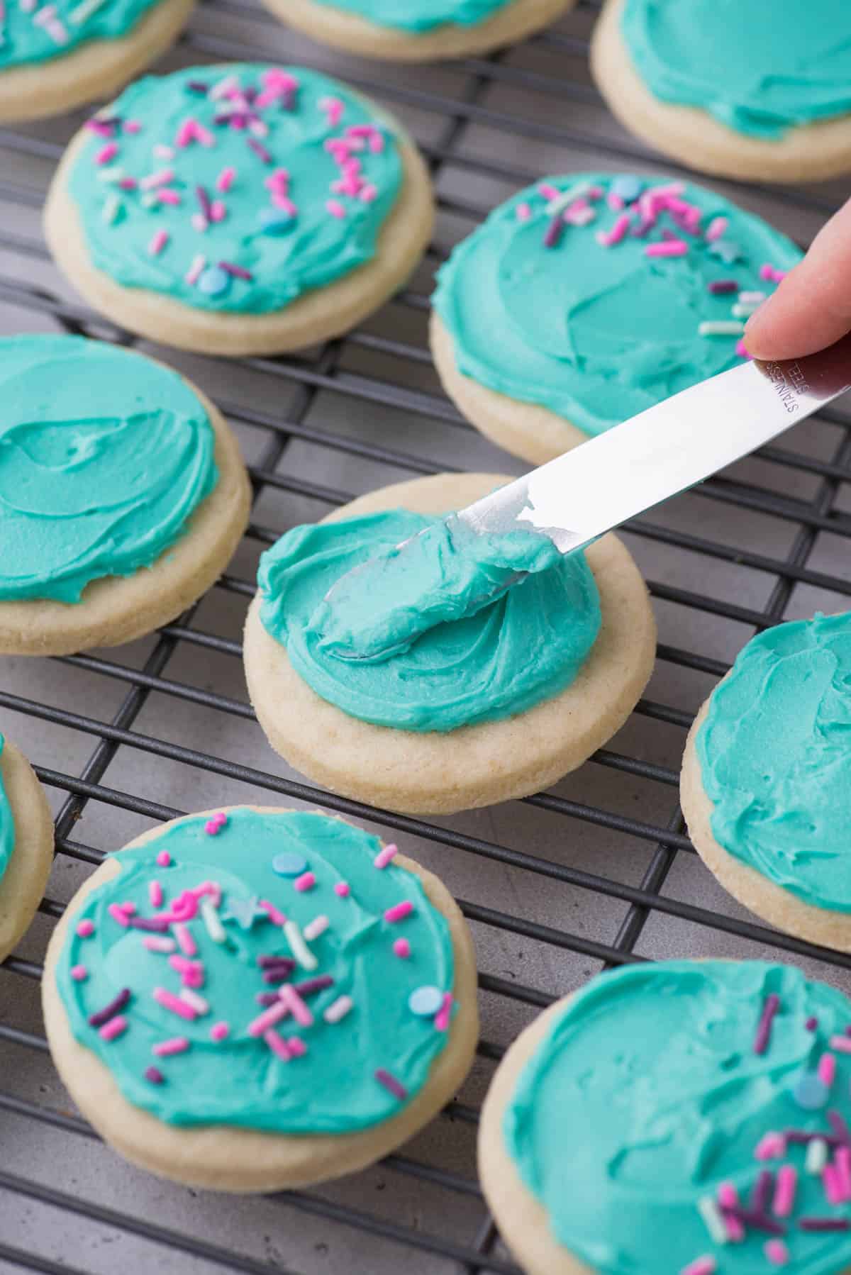 light teal frosting being spread onto a sugar cookie that is on a wire rack in rows with other cookies that have already been frosted and topped with sprinkles