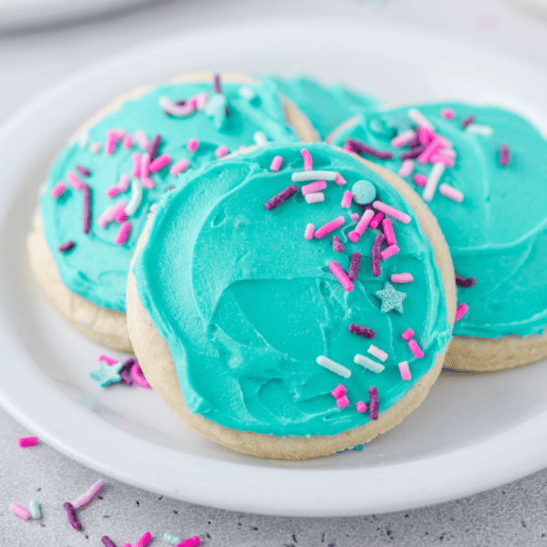 three sugar cookies on a white plate topped with a light teal sugar cookie frosting and sprinkles