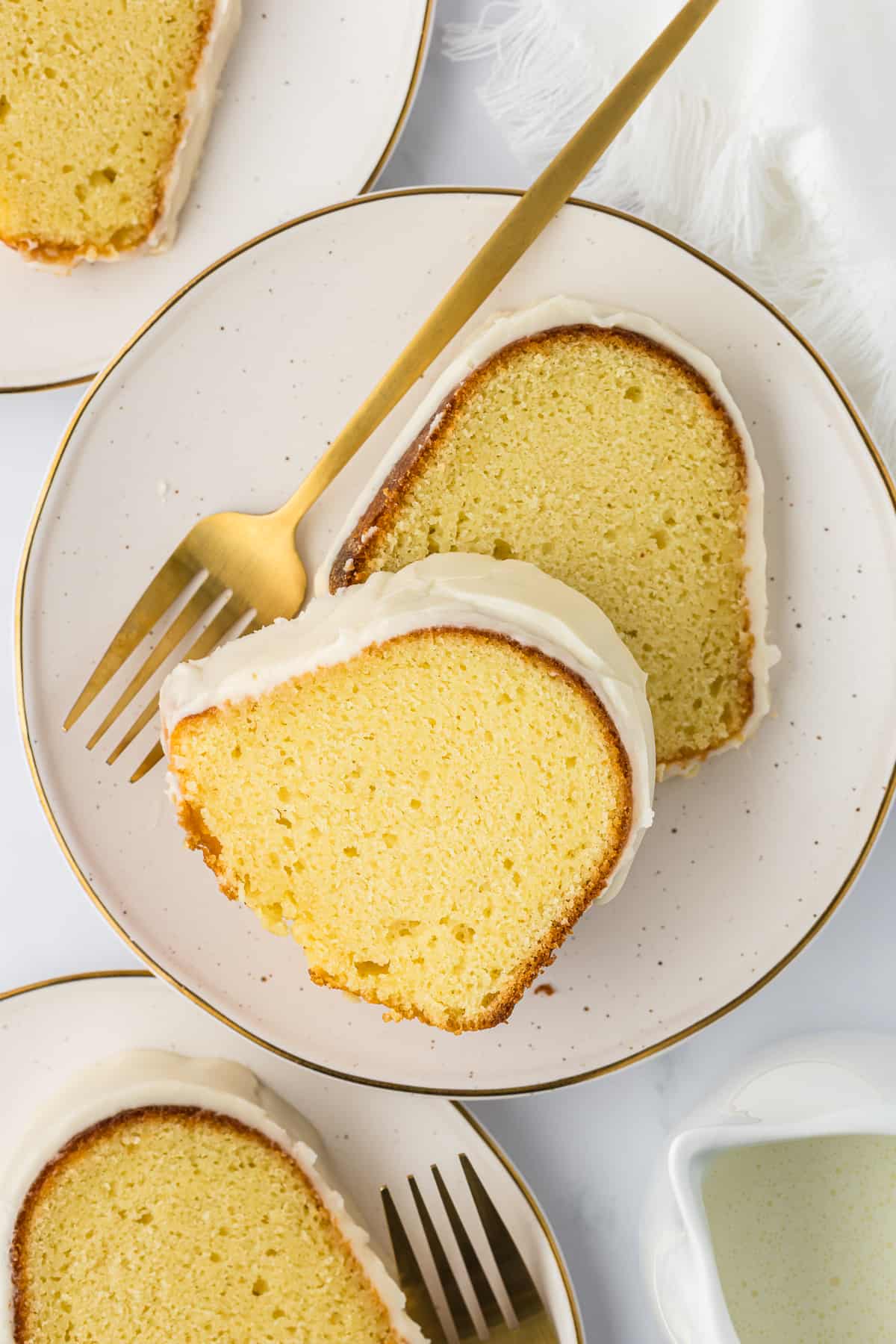 overhead view of two slices of vanilla bundt cake on a small white plate with a fork, with two more plates with a slice of cake beside it