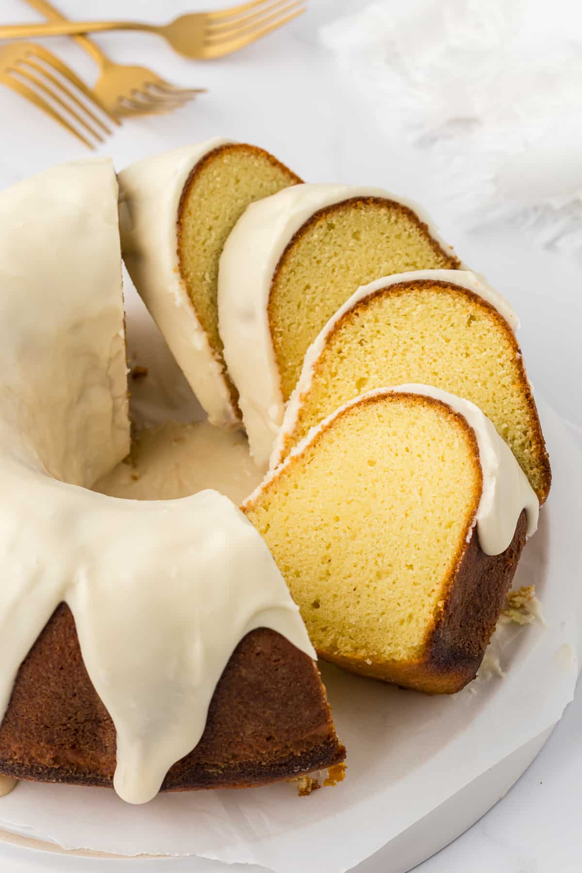 a vanilla bundt cake on a white surface with half the cake in slices and half whole, with three forks laying beside the cake