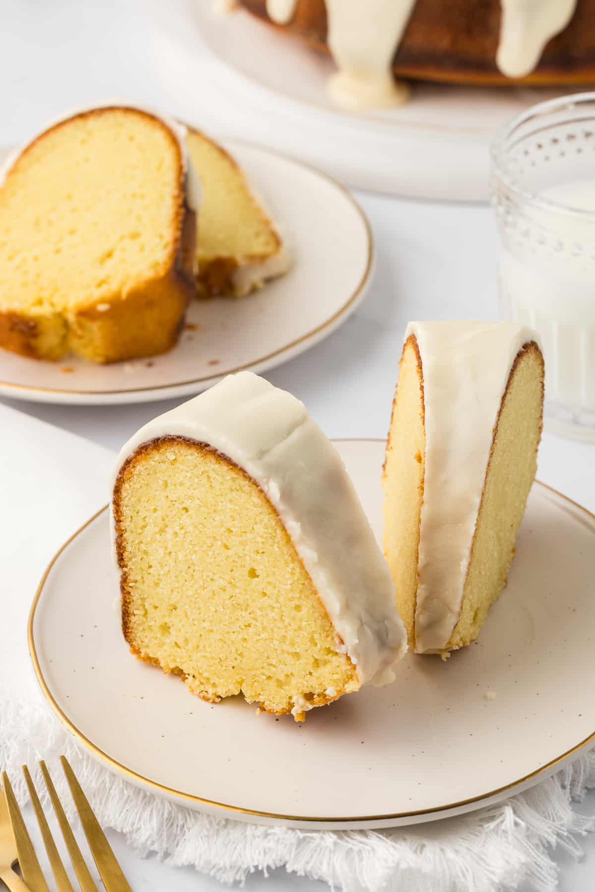 two slices of vanilla bundt cake on a small white plate with a fork beside it, another plate with slices behind it, and a small glass of milk
