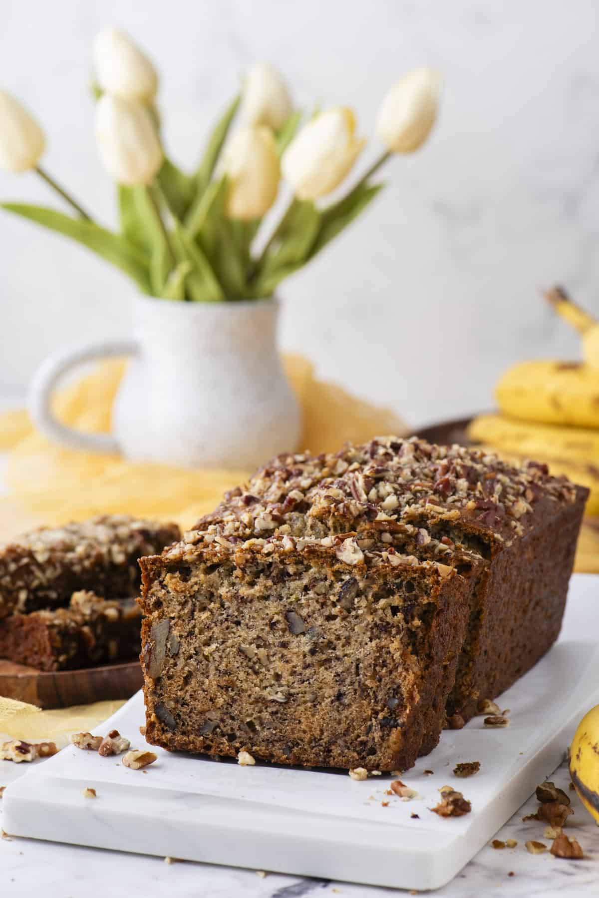 a loaf of banana nut bread on a white cutting board with chopped pecans scattered around, two slices on a wood plate, whole bananas, a yellow cloth and white vase of tulips in the background