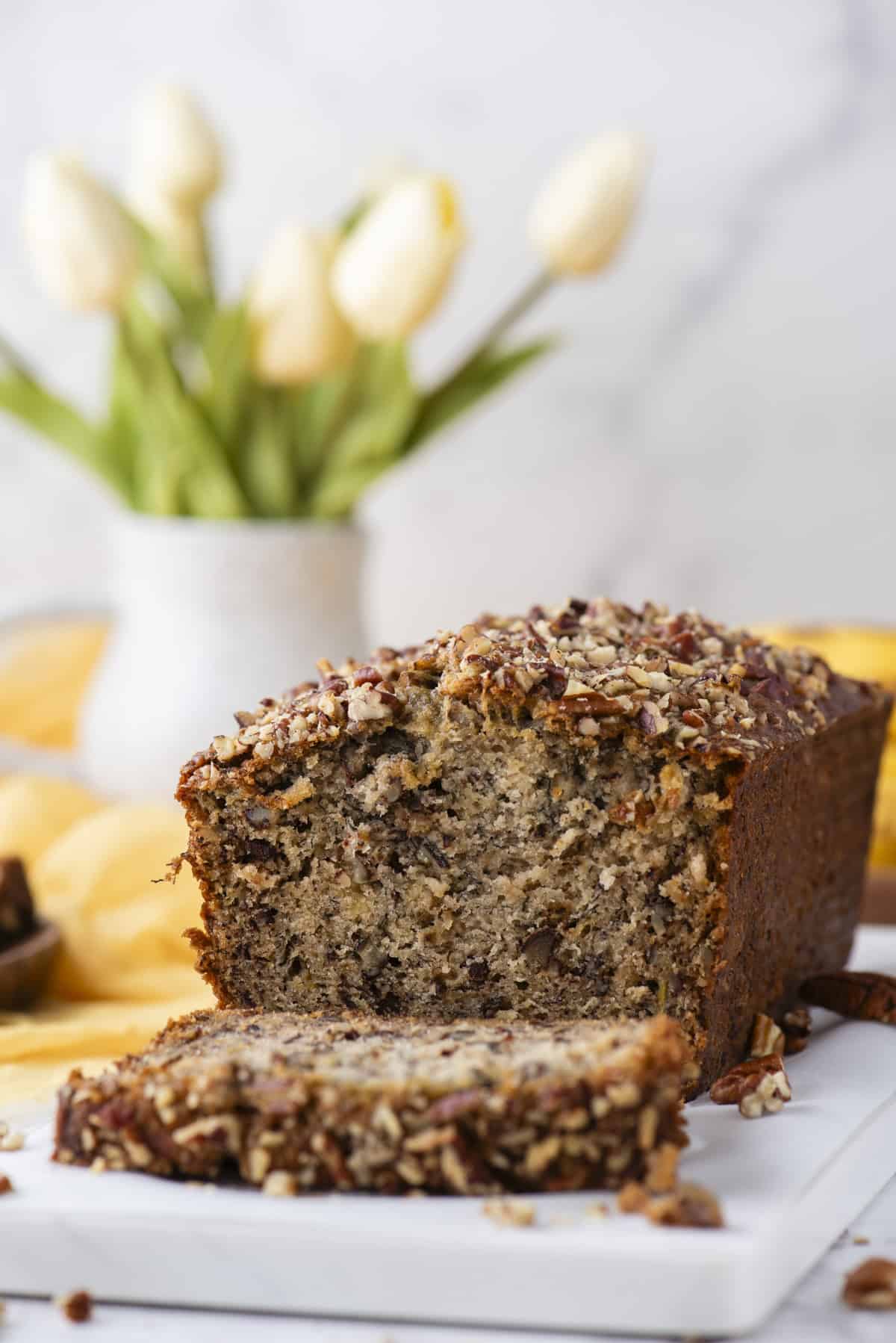 a loaf of banana nut bread on a white cutting board with one slice cut laying in the front and a yellow cloth and white vase of tulips in the background