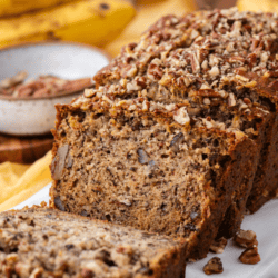 sliced loaf of banana nut bread on a white cutting board with a bowl of nuts in the background