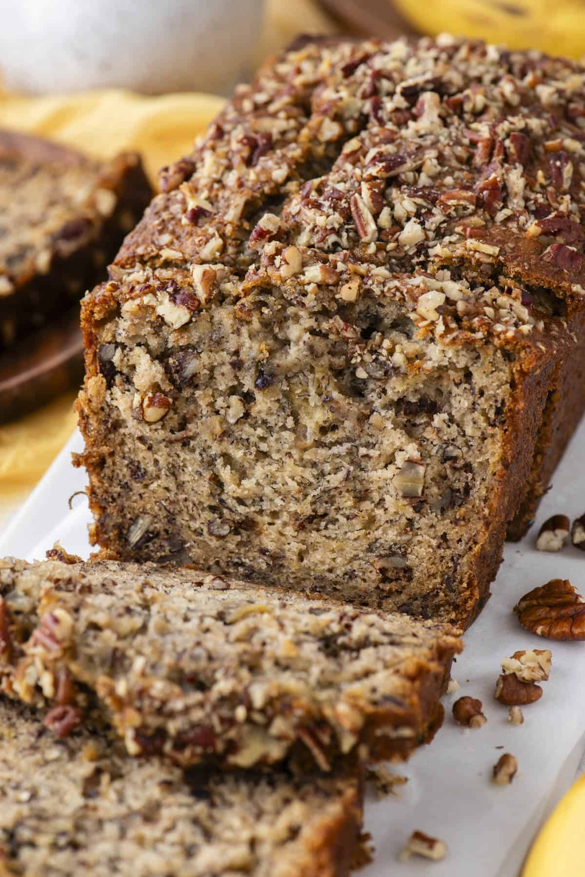 close up of a loaf of banana nut bread on a white cutting board that has been sliced with some slices falling forward on the cutting board and chopped pecans scattered around