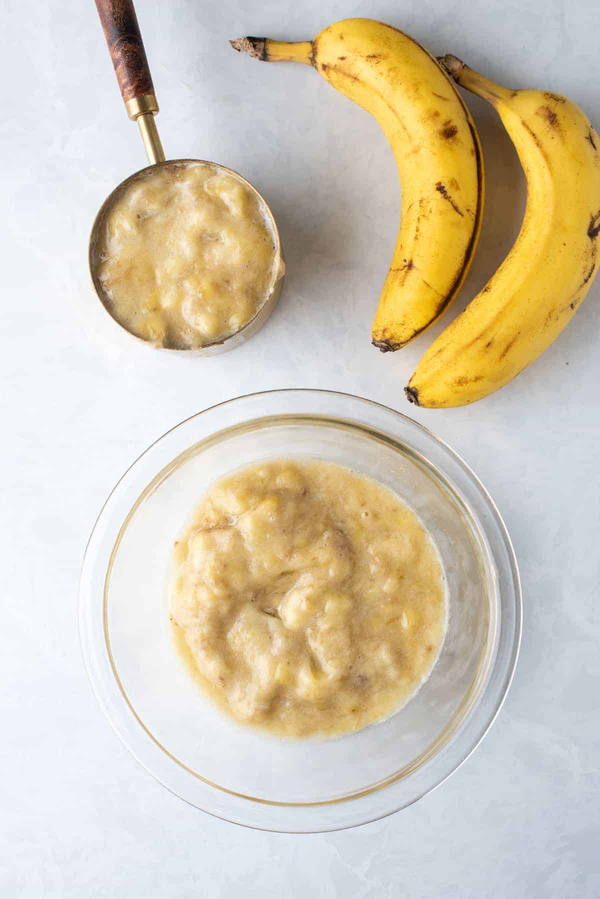overhead view of two whole bananas, a measuring cup full of mashed banana and clear glass bowl full of mashed banana