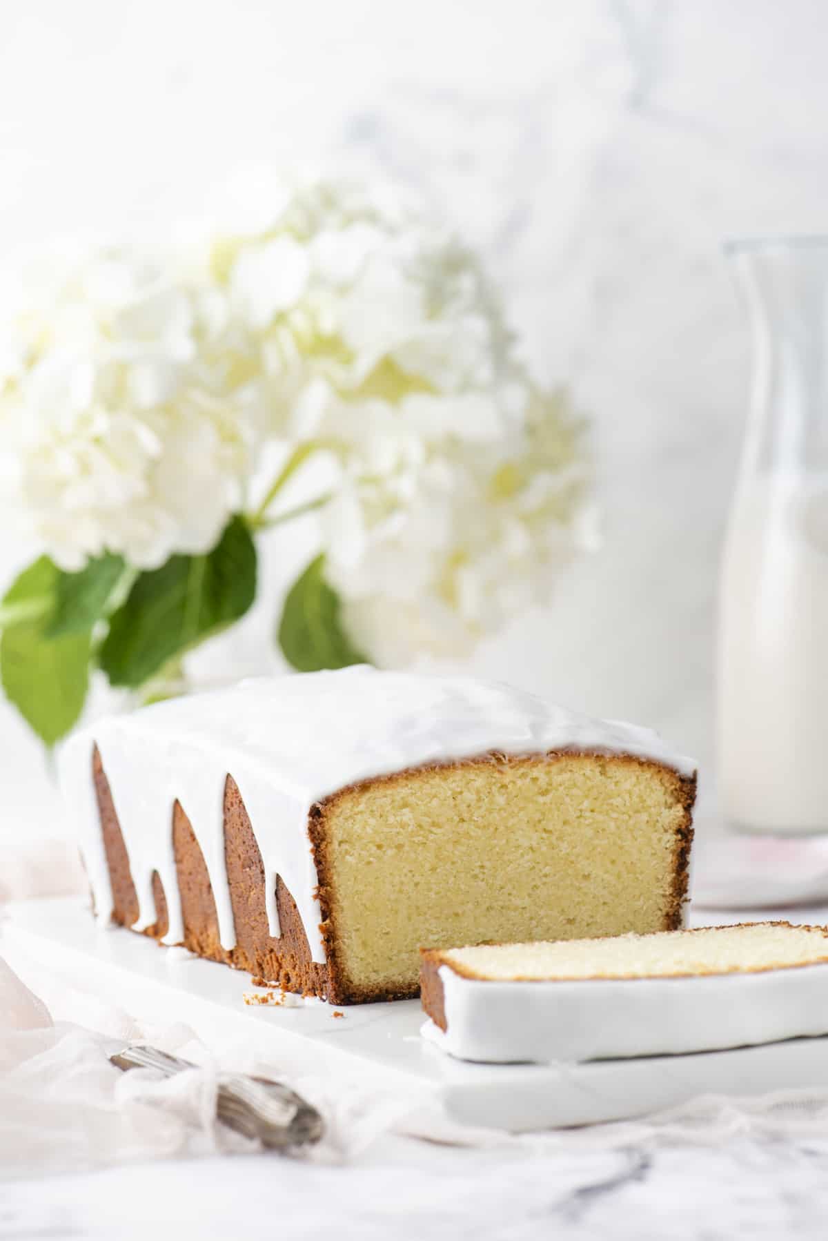 a cream cheese pound cake with one slice cut off laying on its side on a white cutting board with white flowers with green leaves in the background