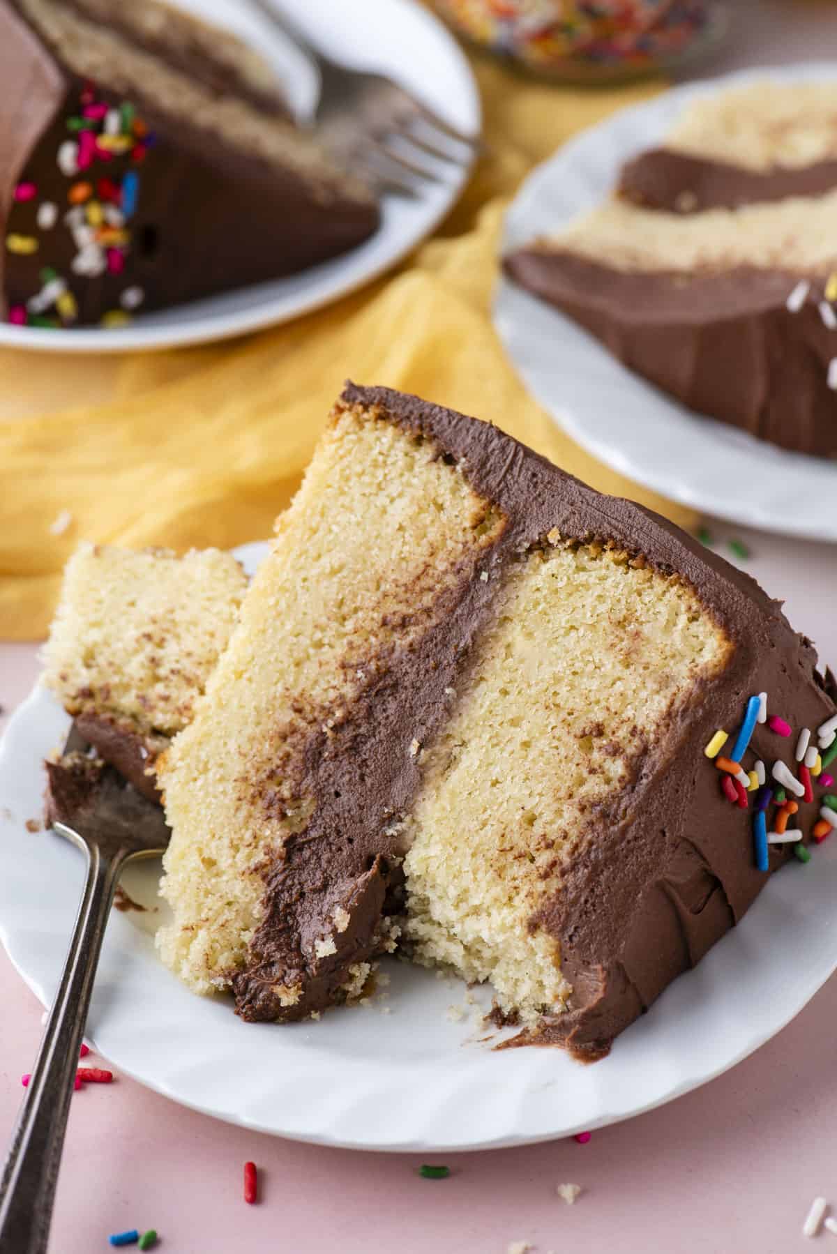 a slice of two layer yellow cake with chocolate frosting on a white plate, with a fork leaning on it that has one bite of cake on it, sitting on a light pink surface with more slices on plates in the background and sprinkles scattered around