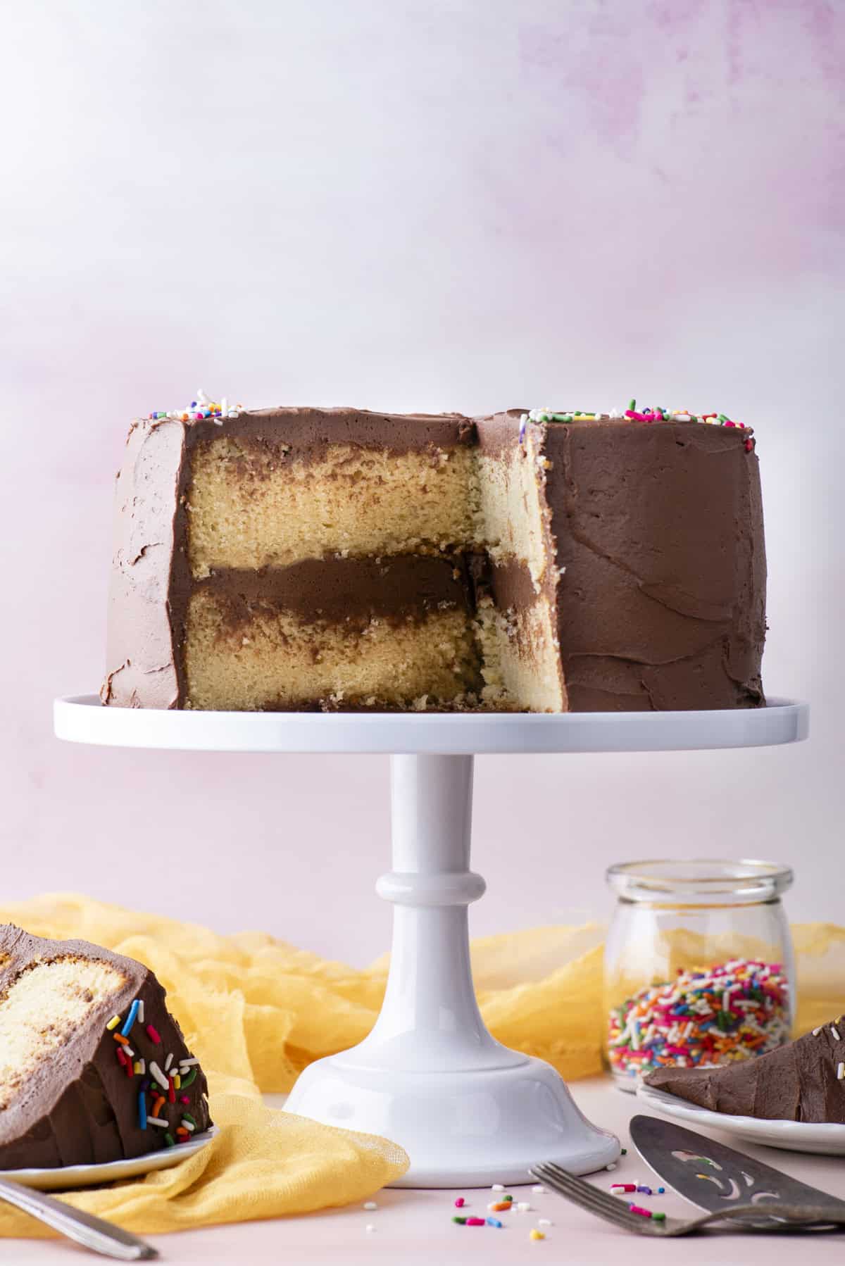 a two layer cake with chocolate frosting and rainbow sprinkles on top, on a white cake stand, with a yellow cloth, two plates with slices of cake, a fork and metal serving spatula, and a clear glass jar of rainbox sprinkles beneath it