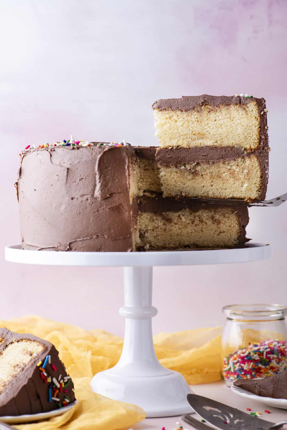 a slice of two layer yellow gluten free cake with chocolate frosting being lifted away from the rest of the cake on a white cake stand, with a slice on a white plate, a fork and metal spatula, a yellow cloth, and a clear class jar of rainbow sprinkles beneath it