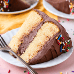 a slice of two layer vanilla cake with chocolate frosting and rainbow sprinkles laying on its side on a small white plate with a fork, sitting on a light pink surface with more slices on plates in the background