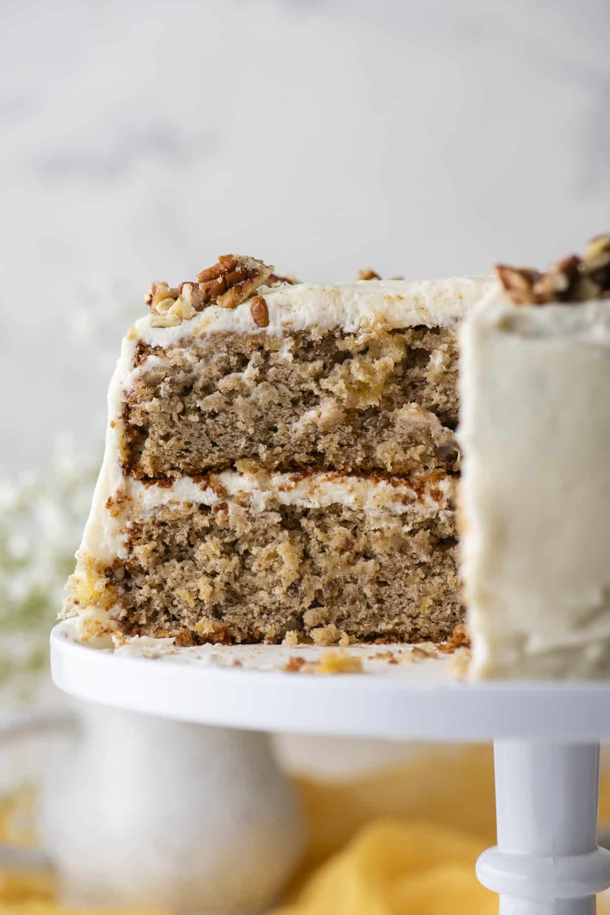 close up of the side view of a hummingbird cake on a white cake stand with one slice missing, exposing the inside of the two layers of cake with cream cheese frosting in between the layers