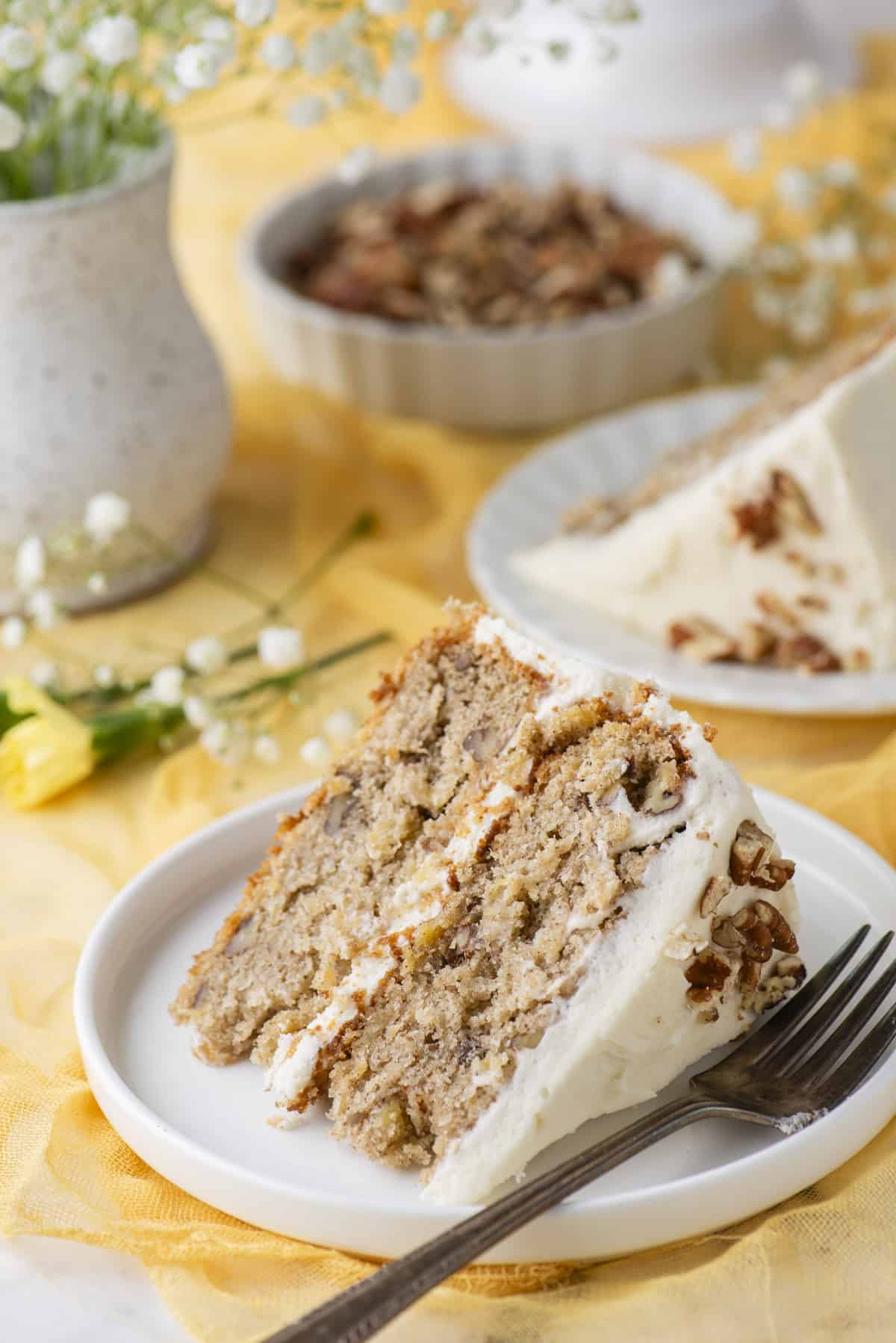 a slice of two-layer hummingbird cake on a white plate with a fork laying beside it, on top of a yellow cloth, with yellow and white flowers and another slice on a plate and a dish of pecans in the background