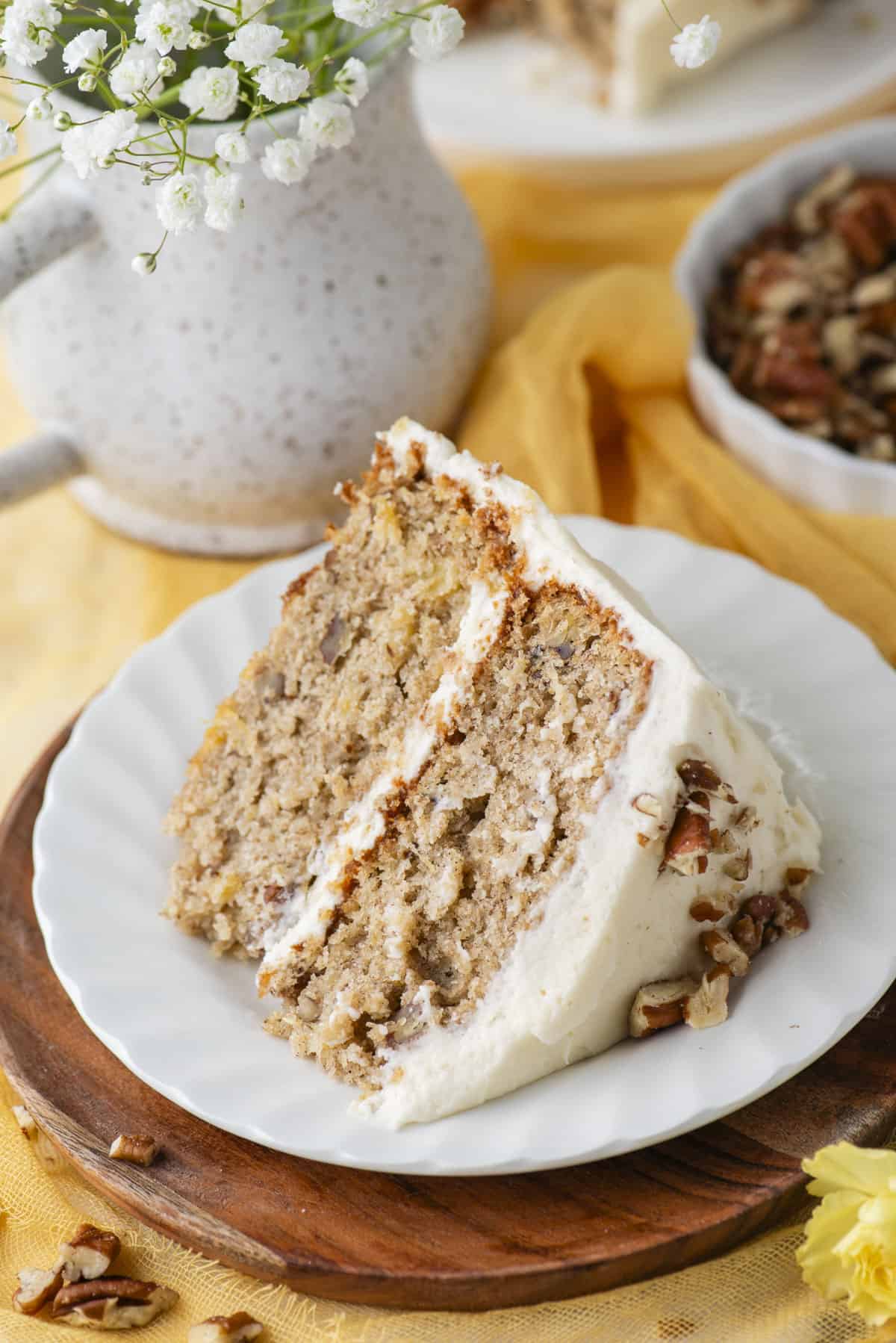 a slice of hummingbird cake on a small white plate on top of a round wood piece beside a dish of pecans and a vase of flowers
