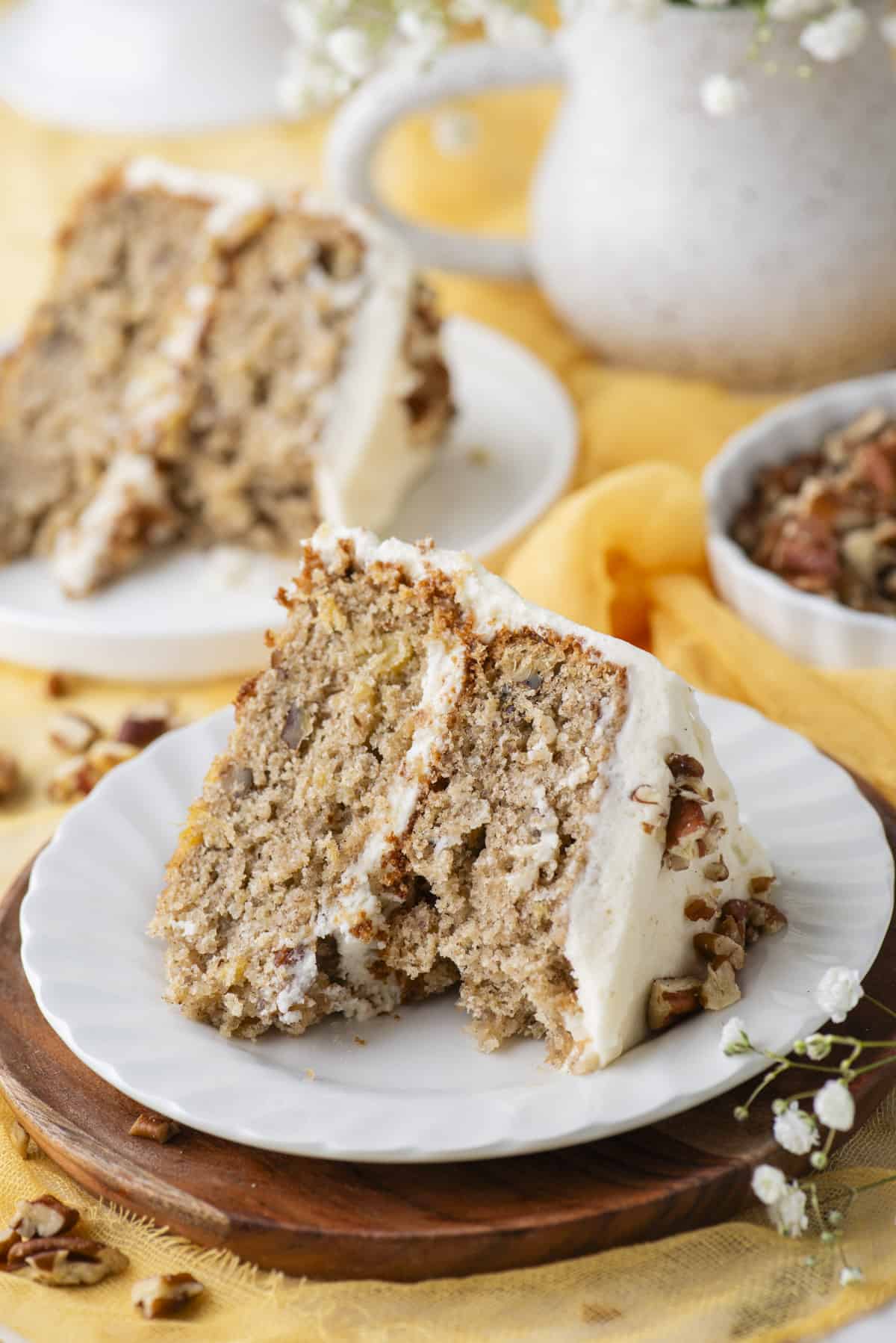 a slice of hummingbird cake on a white plate on top of a wooden circle, with one bite missing from the cake, chopped pecans sprinkled around, some white flowers in the corner and another plate with a slice of cake and a dish of pecans in the background