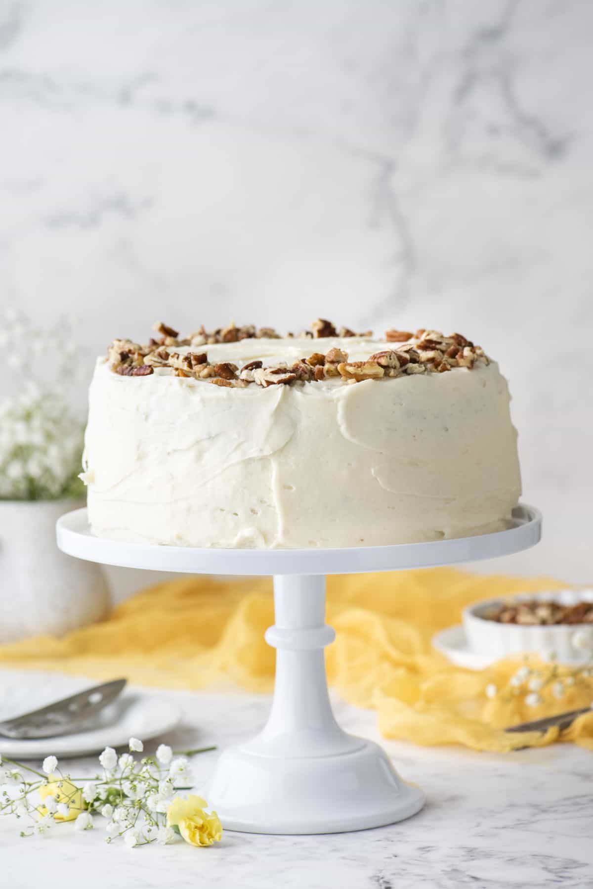 a hummingbird cake covered in cream cheese frosting and topped with chopped pecans on a white cake stand surrounded by white and yellow flowers, a yellow cloth and a bowl of pecans in the background