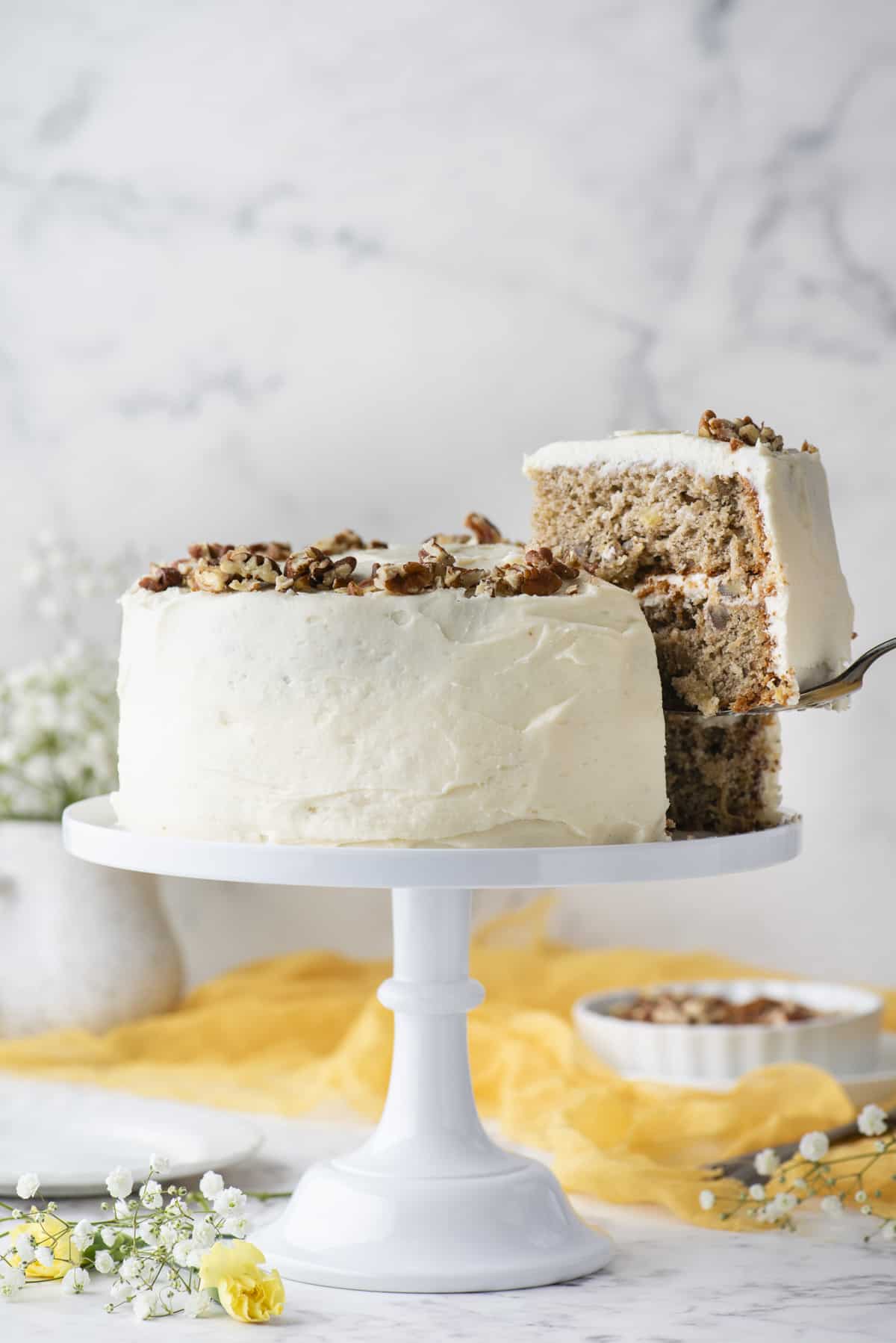 a hummingbird cake on a white cake stand with a piece of the cake being lifted away with a spatula, beneath it white and yellow flowers, a yellow cloth and a white dish of pecans