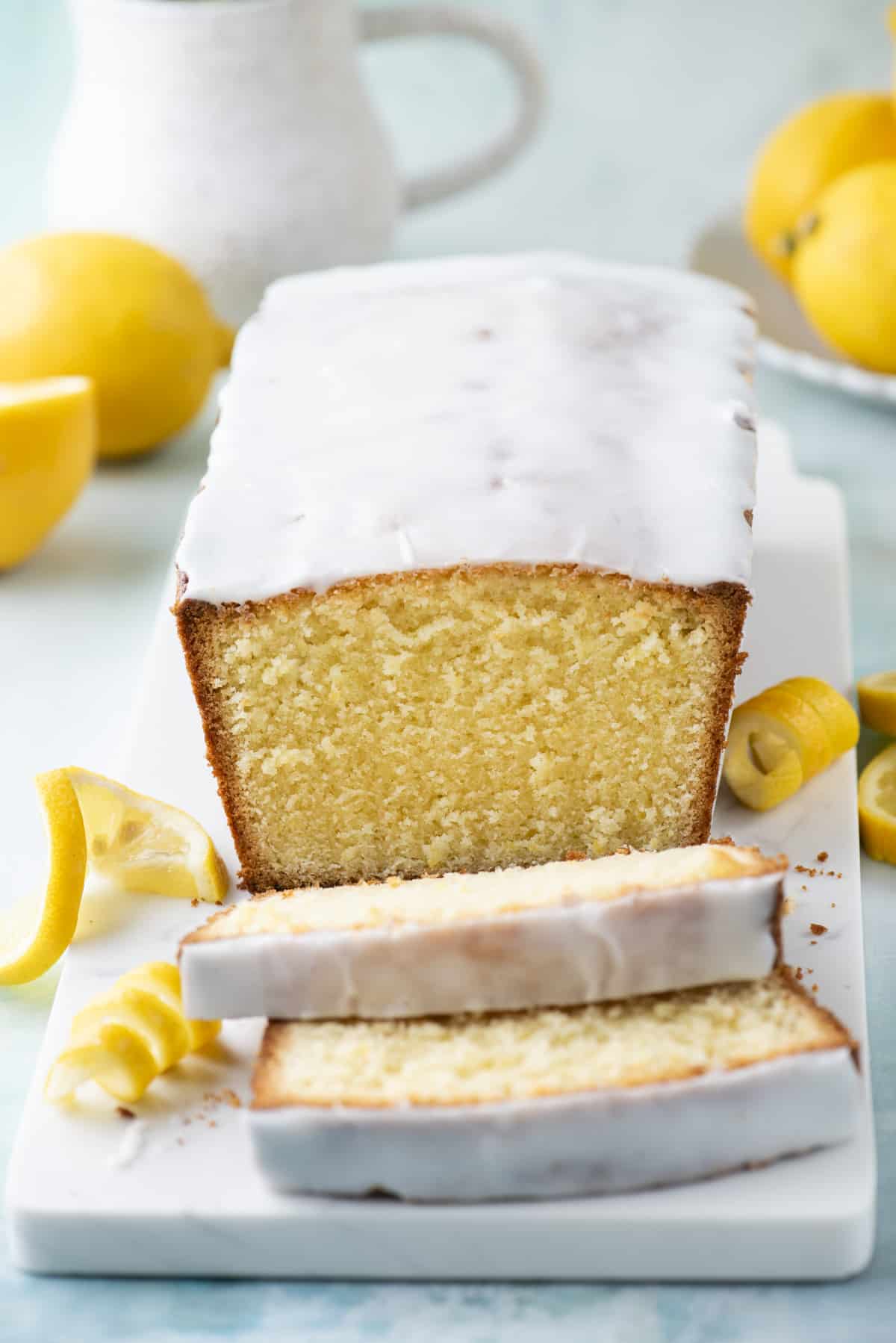 a sliced lemon pound cake on a white cutting board surrounded by fresh lemon slices and lemon peels