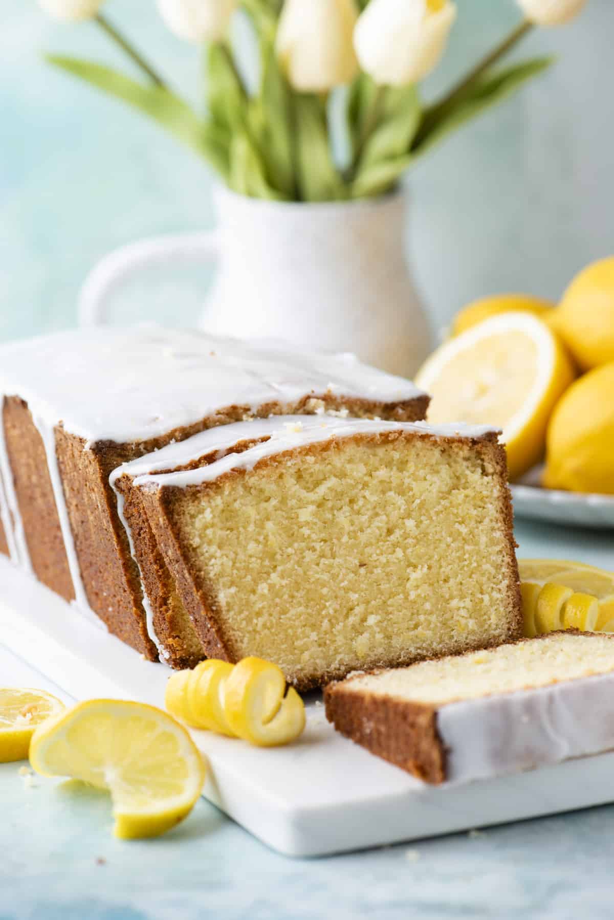 sliced lemon pound cake on a white cutting board surrounded by fresh lemon slices, peels and fresh flowers in the background