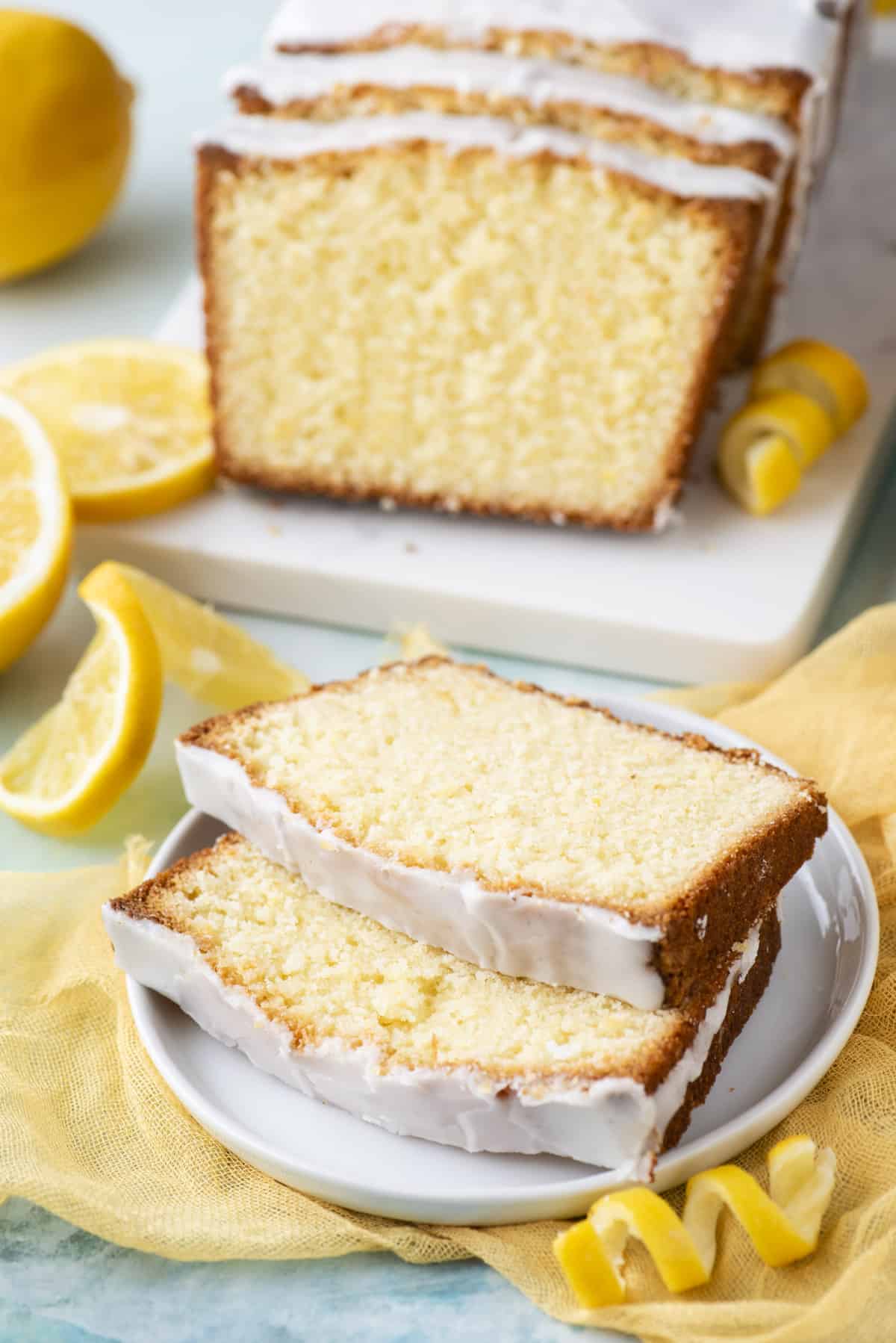 two slices of lemon pound cake on a small white plate with more of the cake and fresh lemon slices and peels in the background
