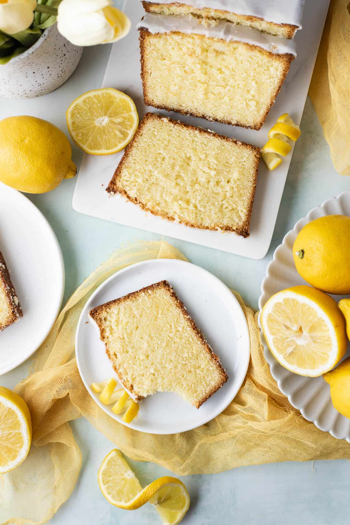 over head view of a lemon pound cake sliced on a white cutting board on a light blue surface surrounded by small white plates with slices of cake on them, fresh whole lemonds, lemon slices and peels and fresh flowers