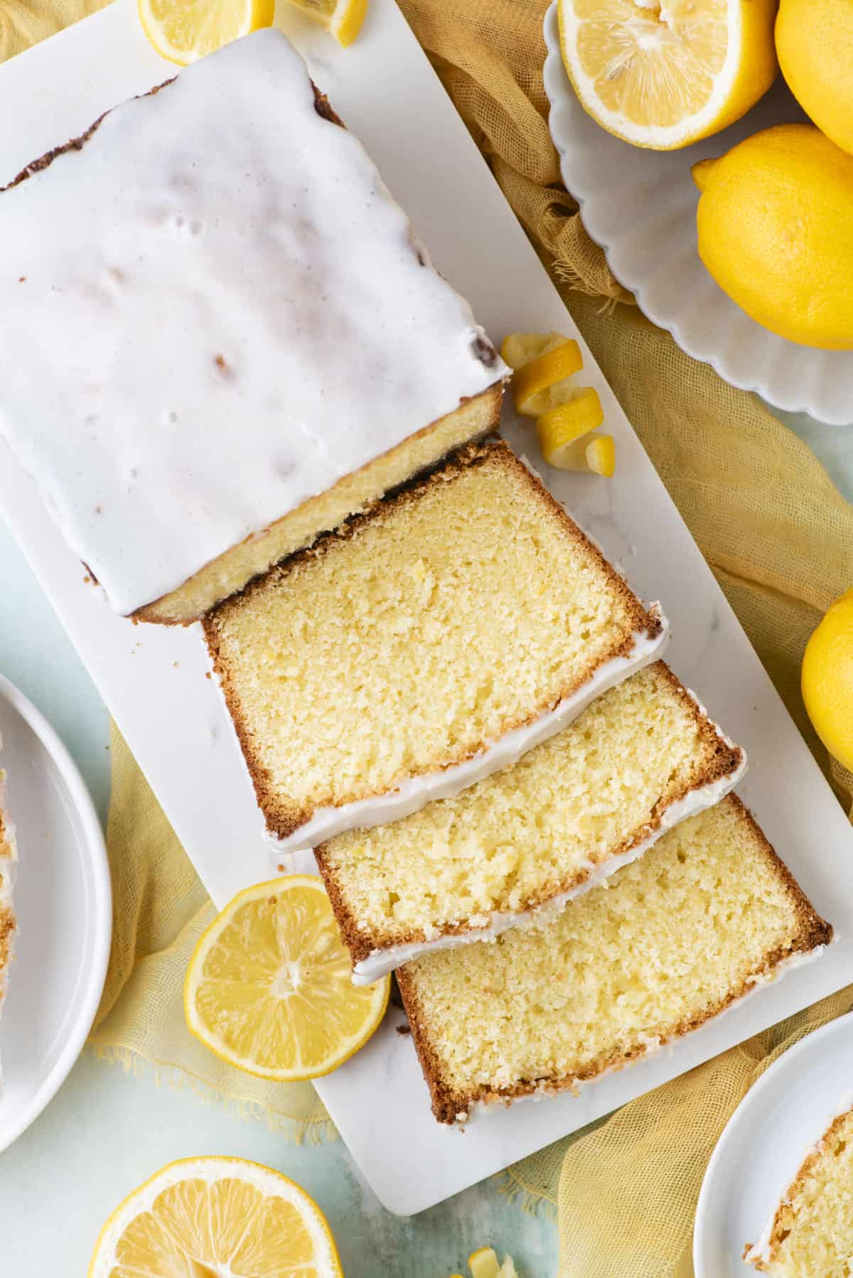 over head view of lemon pound cake, half sliced on a white cutting board surrounded by a bowl of lemons and lemon slices and peels scattered around