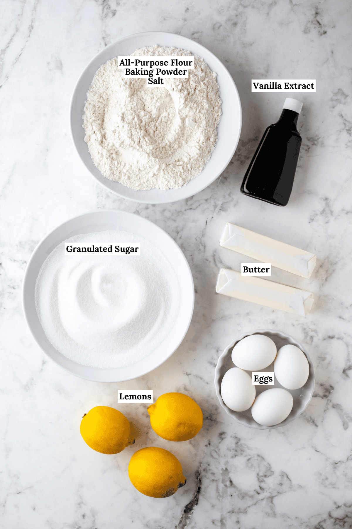 over head view of ingredients for lemon pound cake on a counter top including three whole lemons, 4 eggs, two sticks of butter, a bowl of granulated sugar, a bottle of vanilla extract, and a bowl of all-purpose flour, baking powder and salt
