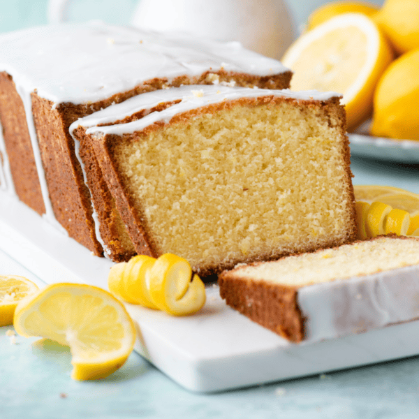 a sliced loaf of lemon pound cake on a white cutting board on a light blue surface with fresh lemon slices and peels scattered around and more whole and sliced lemons in the background