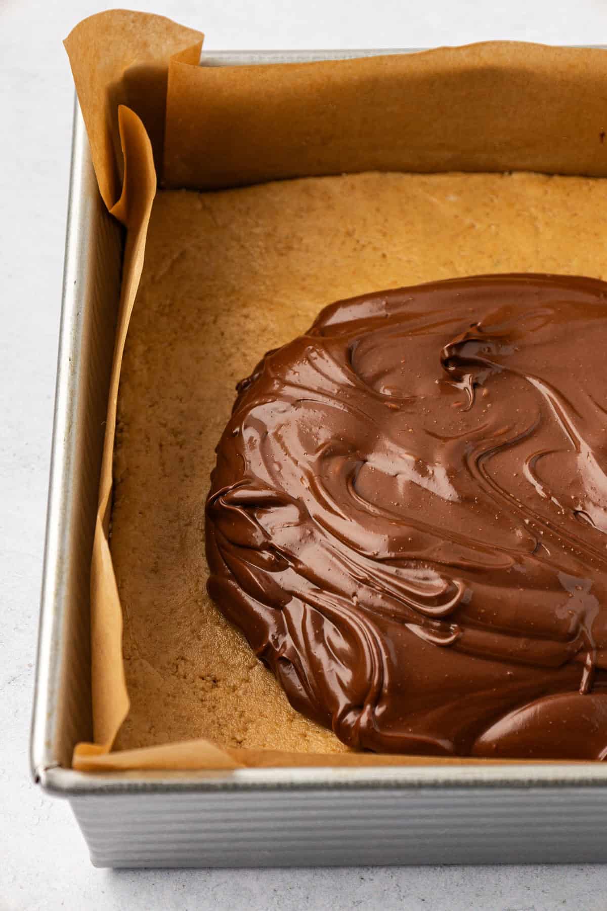 Close up of a square pan lined with brown parchment paper with a smooth peanut butter layer topped with a large dollop of a melted chocolate mixture